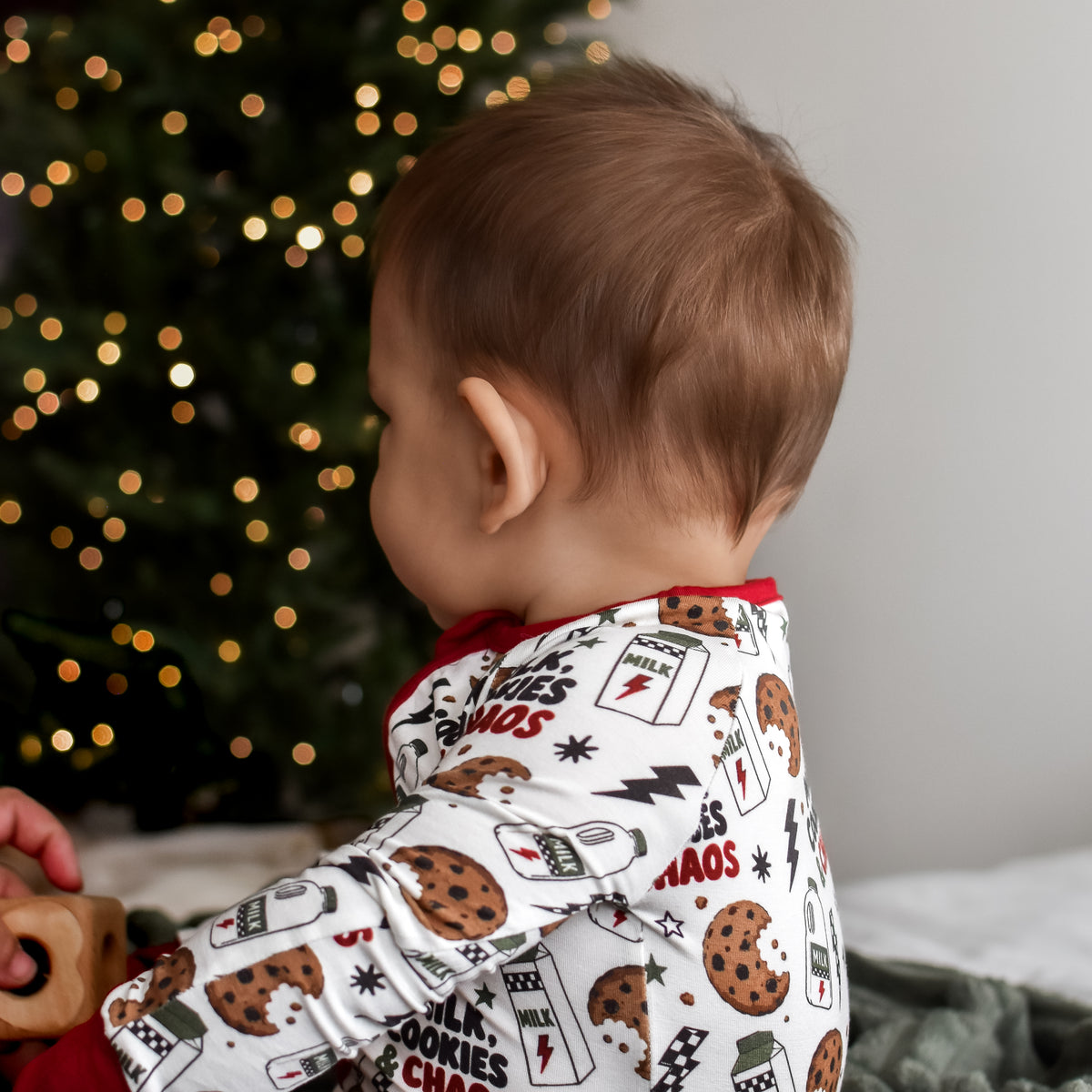 A baby wearing a white Christmas-themed cookie print pajama set, sitting on a gray blanket in front of a glowing Christmas tree.