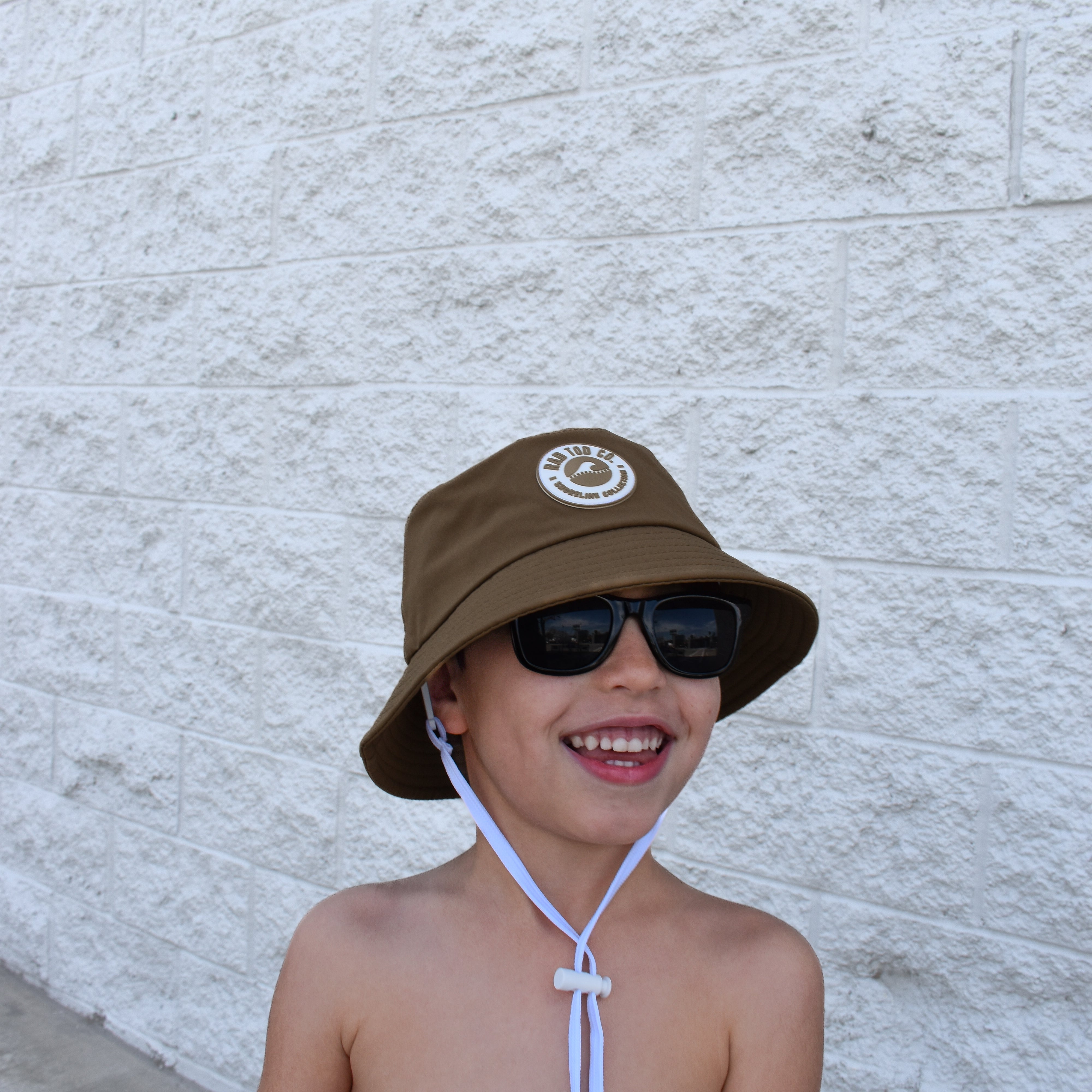Child wearing a brown bucket hat and sunglasses against a light gray wall.