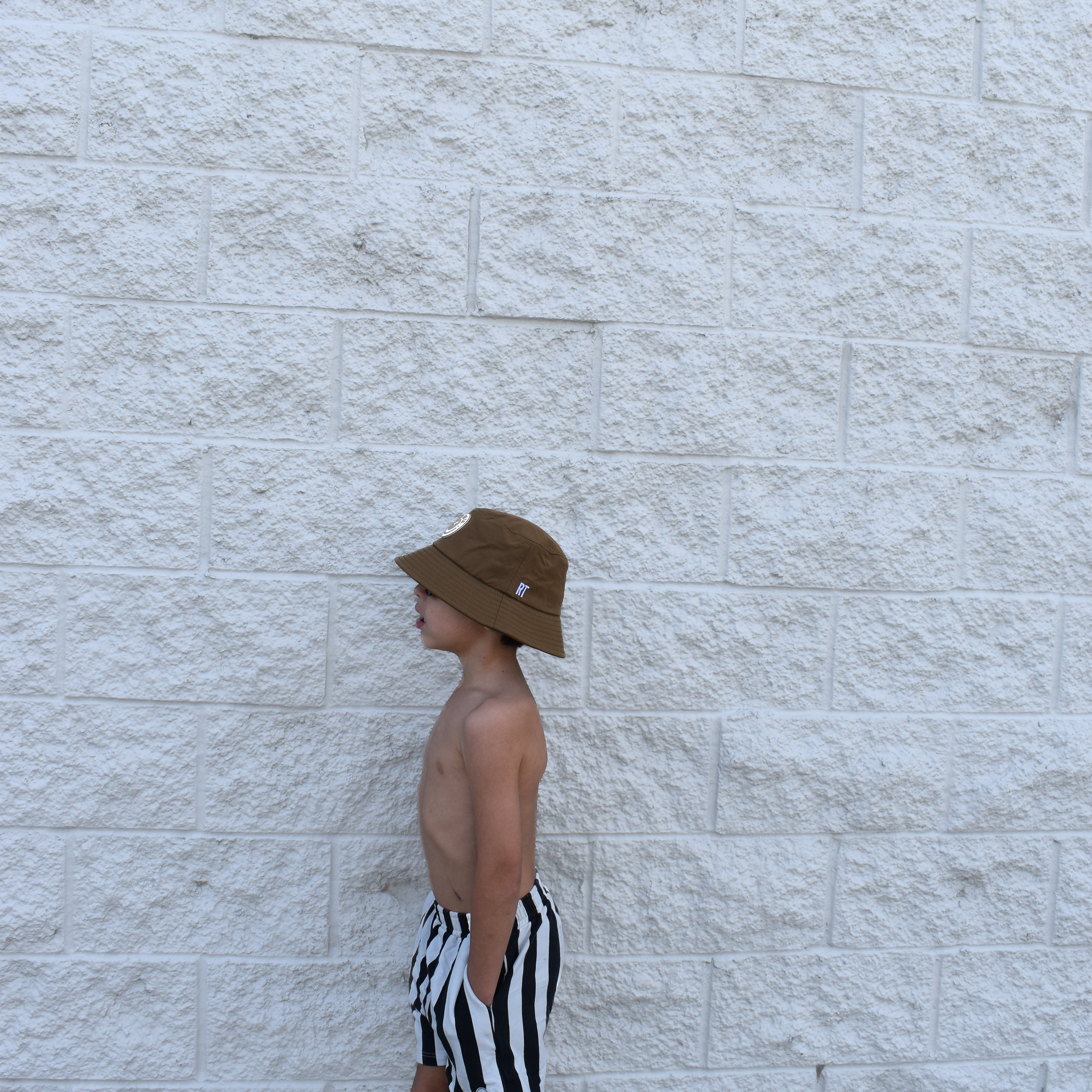 Person wearing a brown bucket hat and black and white striped shorts standing against a light stone wall.