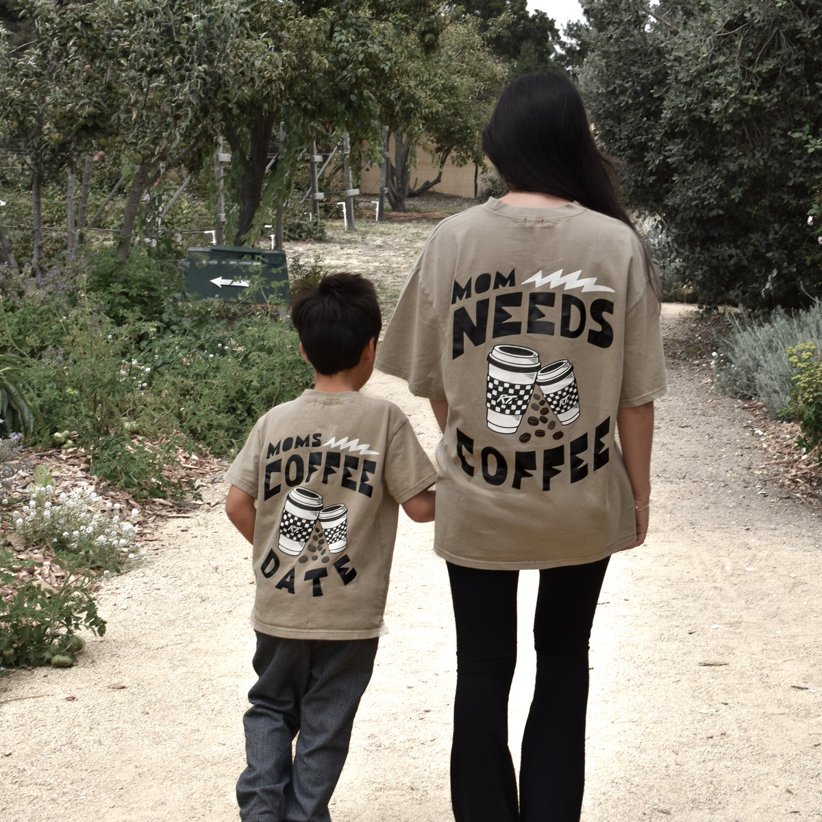 A woman and a young boy walk hand-in-hand on a dirt path, viewed from behind. Both are wearing matching beige graphic t-shirts with black text and coffee cup illustrations.