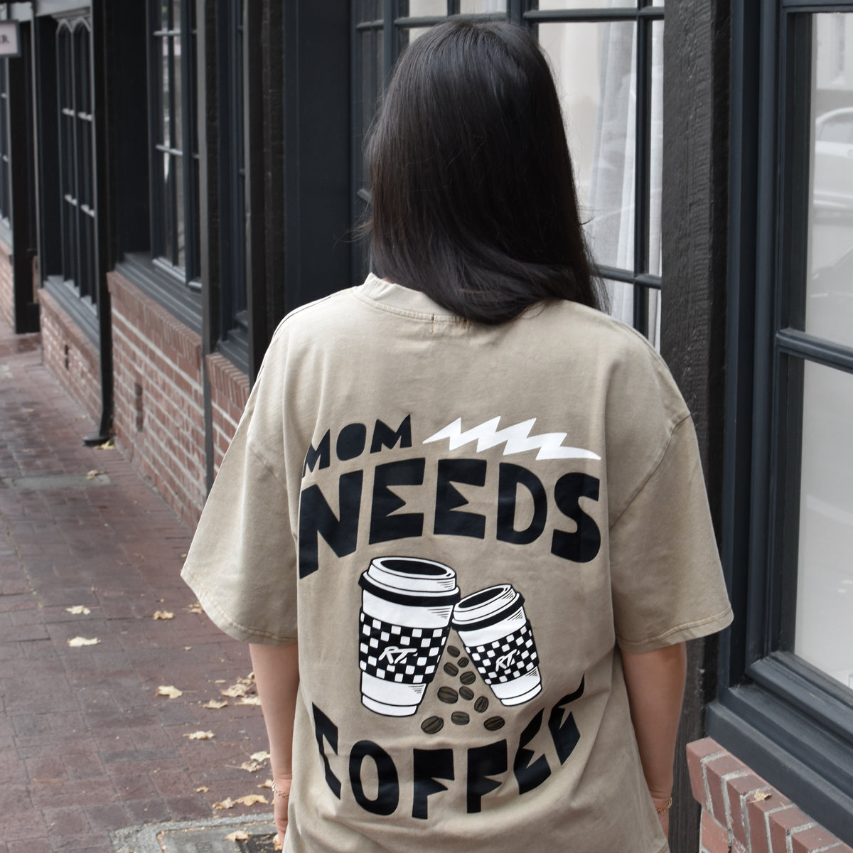 A woman viewed from behind wearing an oversized beige graphic t-shirt with the text 'MOM NEEDS COFFEE' and coffee cup illustrations on the back, standing on a brick sidewalk next to a building with black-framed windows.