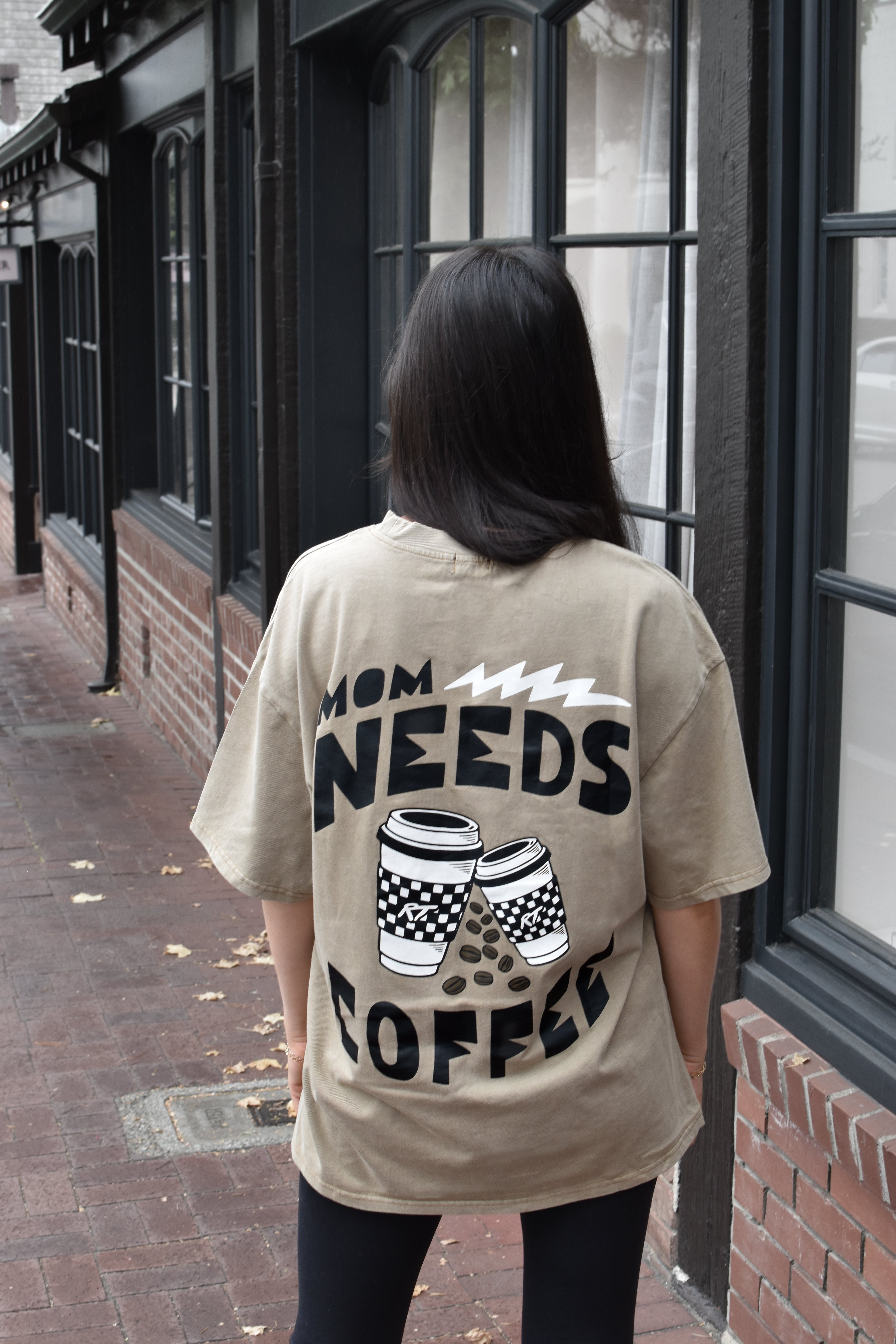 A woman viewed from behind wearing an oversized beige graphic t-shirt with the text 'MOM NEEDS COFFEE' and coffee cup illustrations on the back, standing on a brick sidewalk next to a building with black-framed windows.