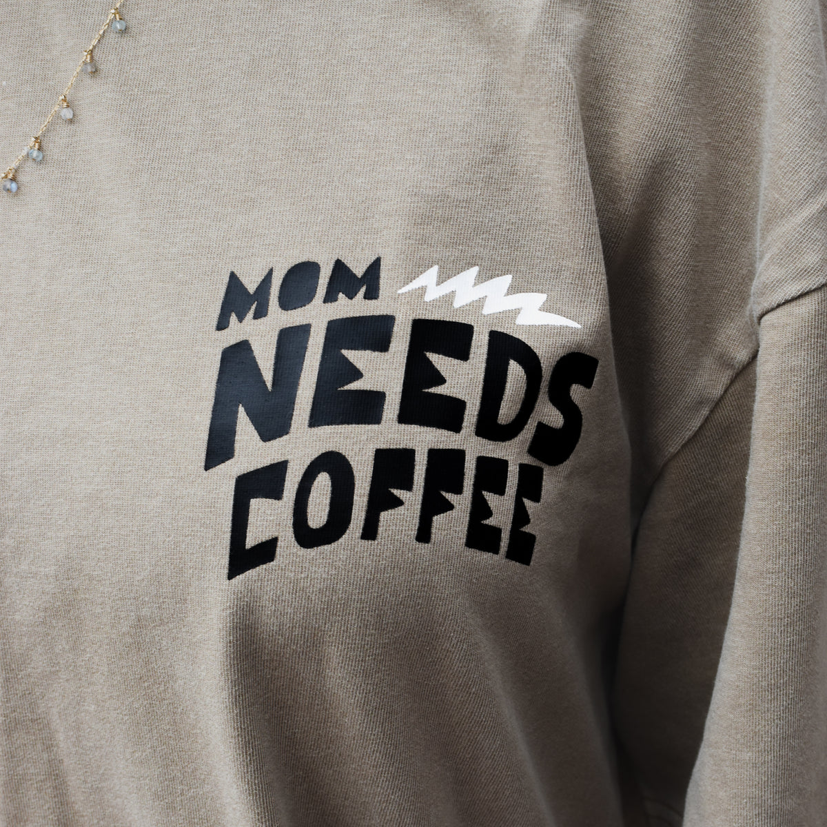 Close-up of a person wearing beige t-shirt with a black and white graphic that reads 'MOM NEEDS COFFEE'.