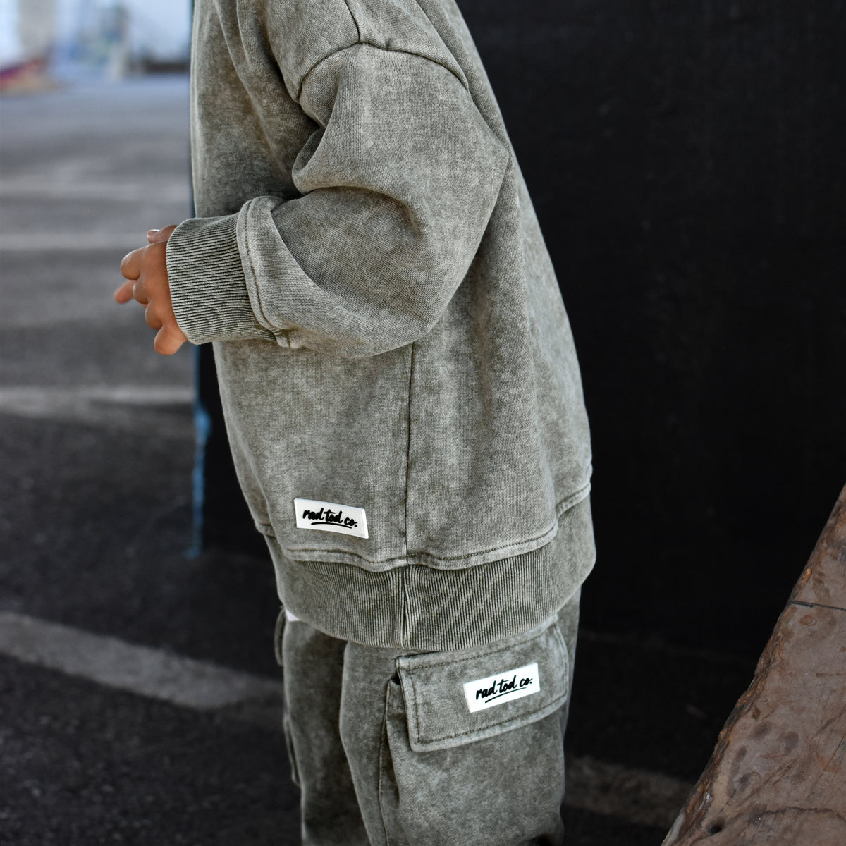 A child wearing a matching moss washed sweatshirt and jogger pants set with small white brand patches, standing outdoors against a dark wall.