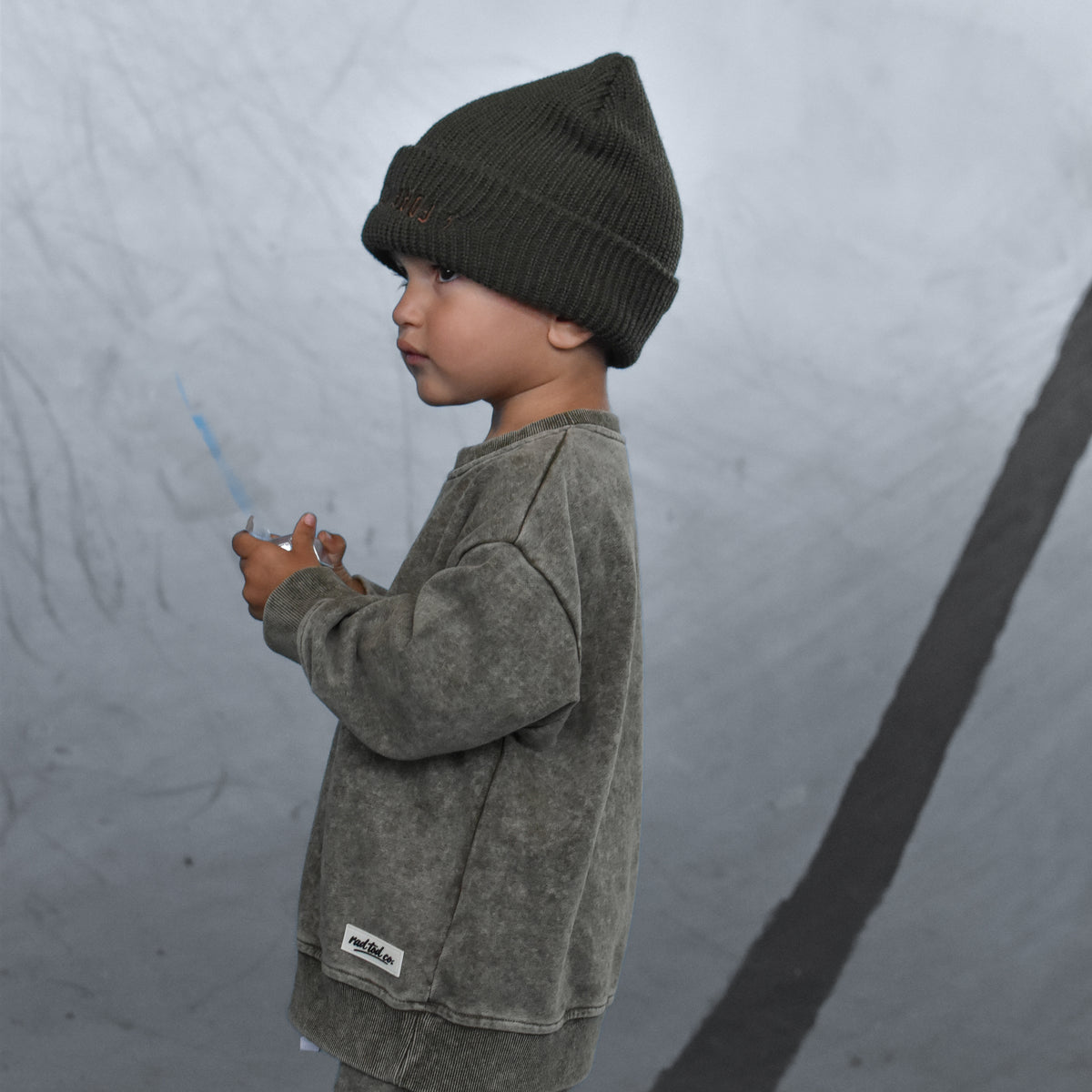 A child standing in profile wearing a dark olive green beanie, a matching washed green sweatshirt, and green cargo pants in a concrete bowl.