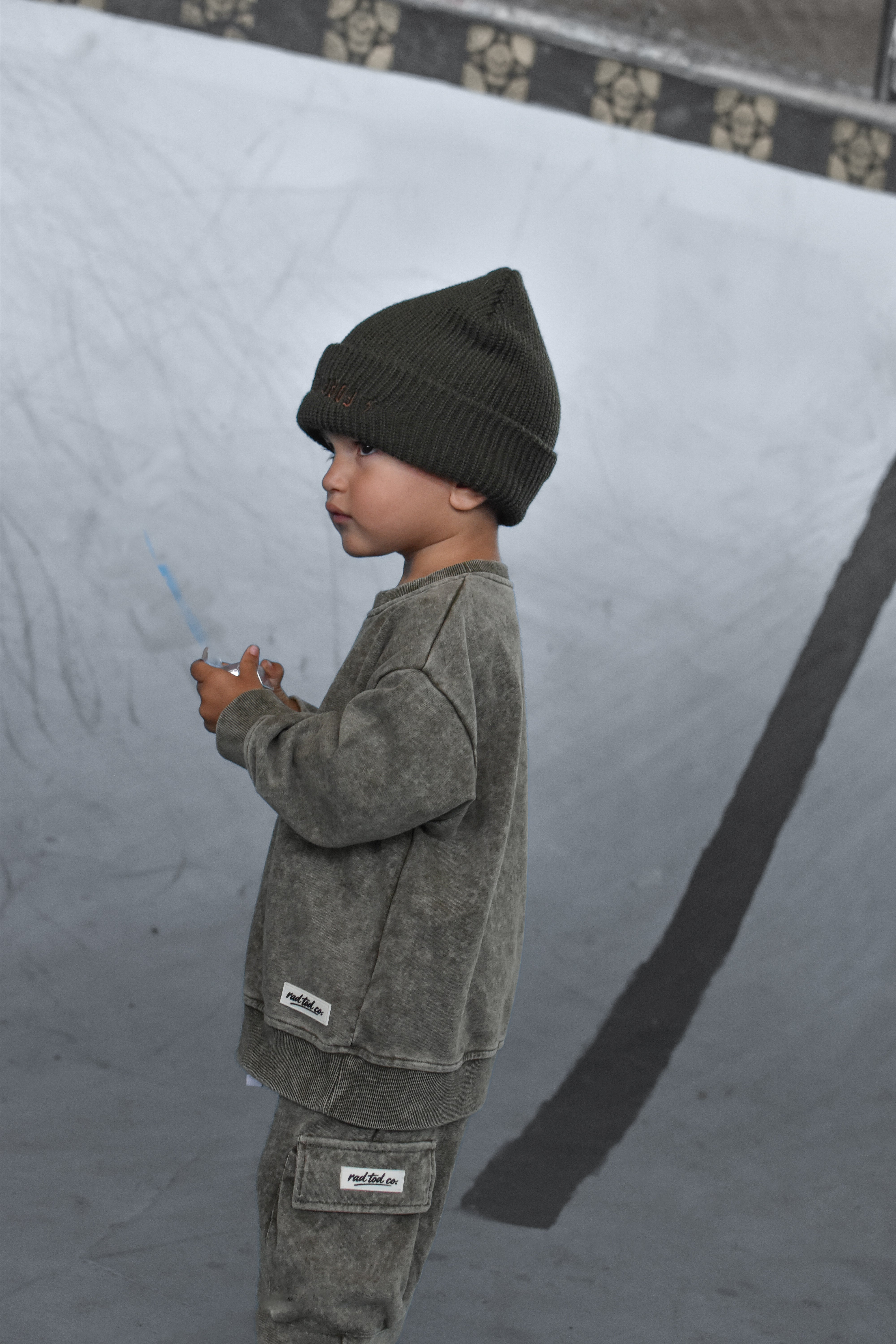 A child standing in profile wearing a dark olive green beanie, a matching washed green sweatshirt, and green cargo pants in a concrete bowl.