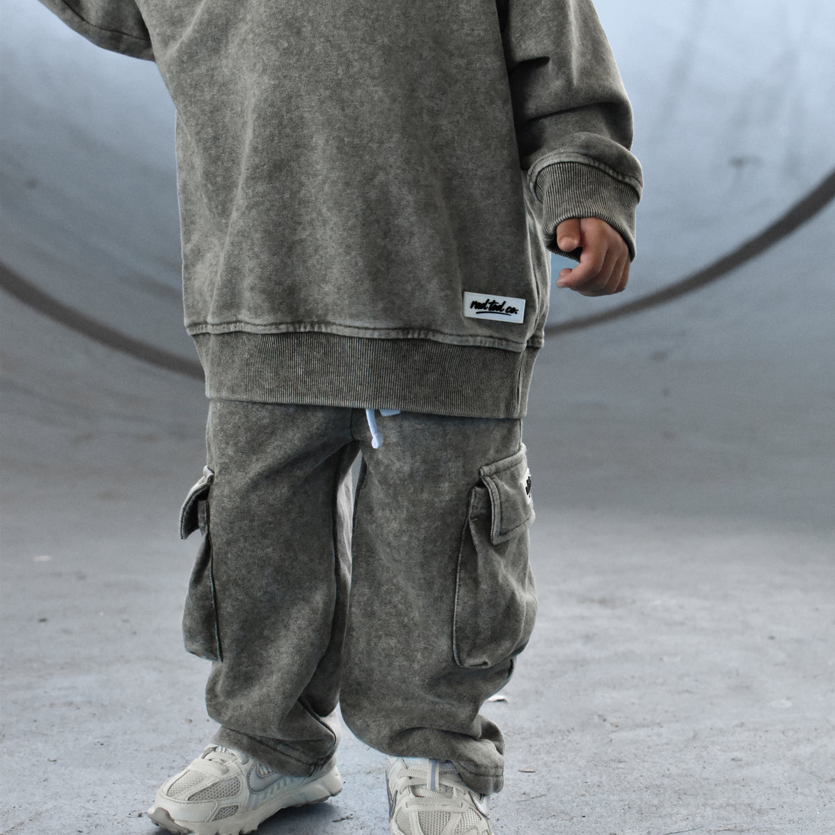 A child wearing a matching washed gmoss sweatshirt and cargo pants set, standing in a concrete skatepark environment.