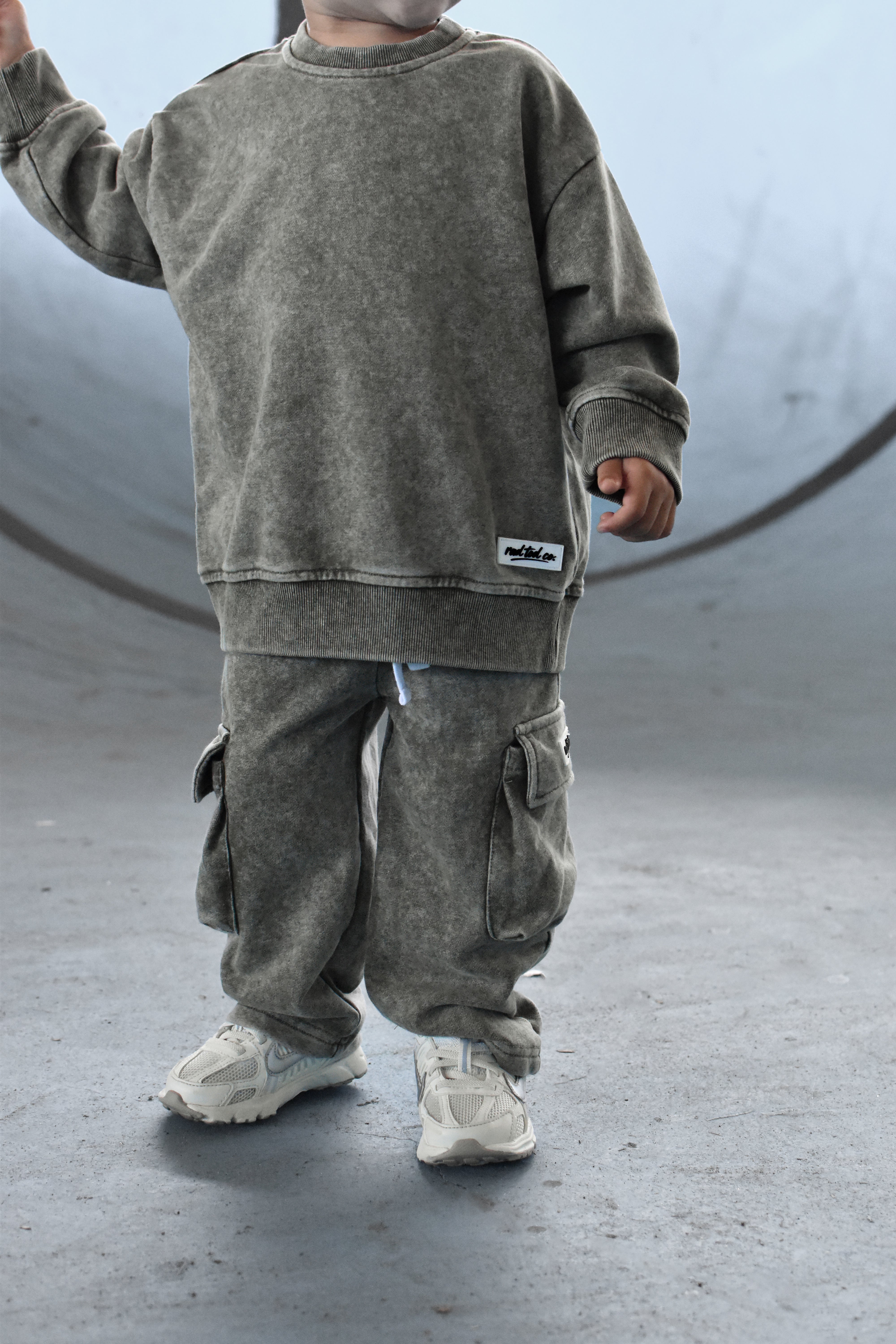 A child wearing a matching washed gmoss sweatshirt and cargo pants set, standing in a concrete skatepark environment.