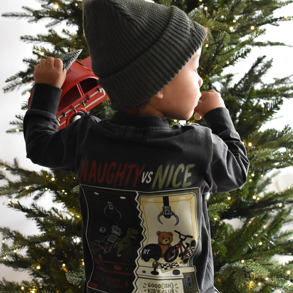 A toddler viewed from behind wearing a dark charcoal gray long-sleeve graphic shirt and a dark knit beanie, standing in front of a decorated Christmas tree.