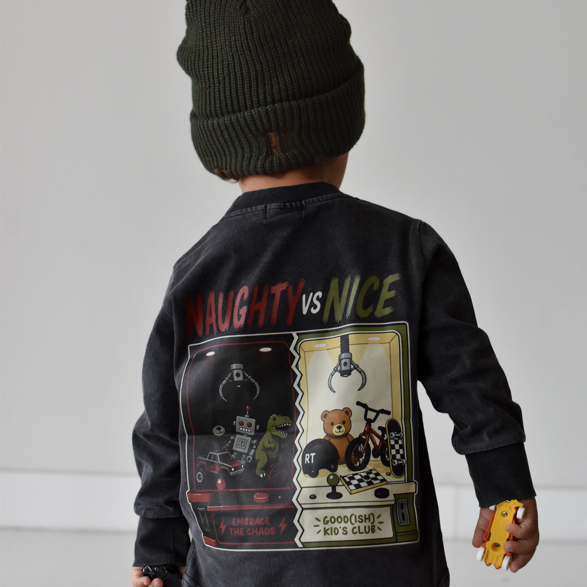 A child shown from the back wearing a dark charcoal long-sleeve graphic jacket with 'NAUGHTY vs NICE' text and a large retro game-style print, paired with a dark green beanie and beige pants against a light gray studio background.