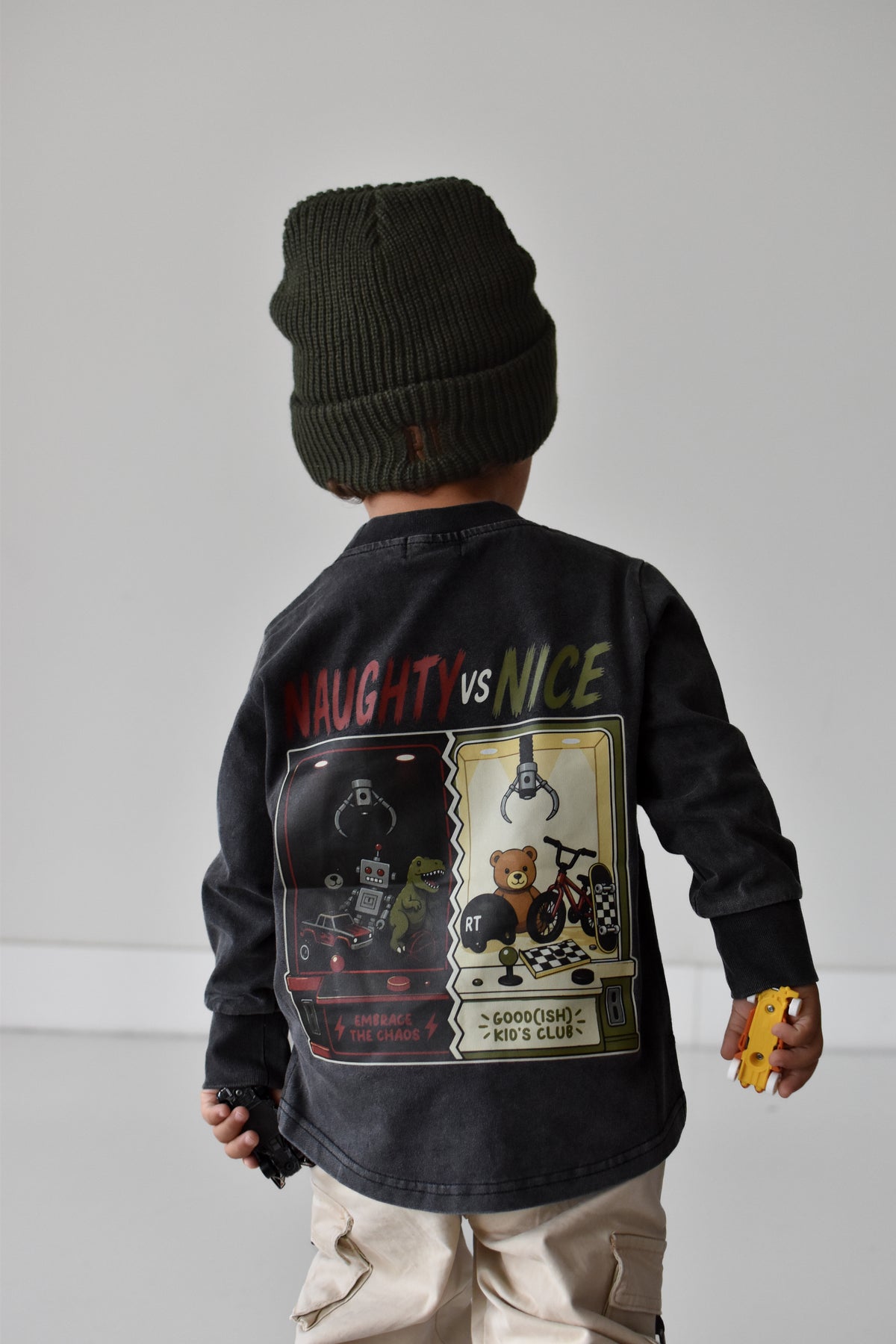 A child shown from the back wearing a dark charcoal long-sleeve graphic jacket with 'NAUGHTY vs NICE' text and a large retro game-style print, paired with a dark green beanie and beige pants against a light gray studio background.