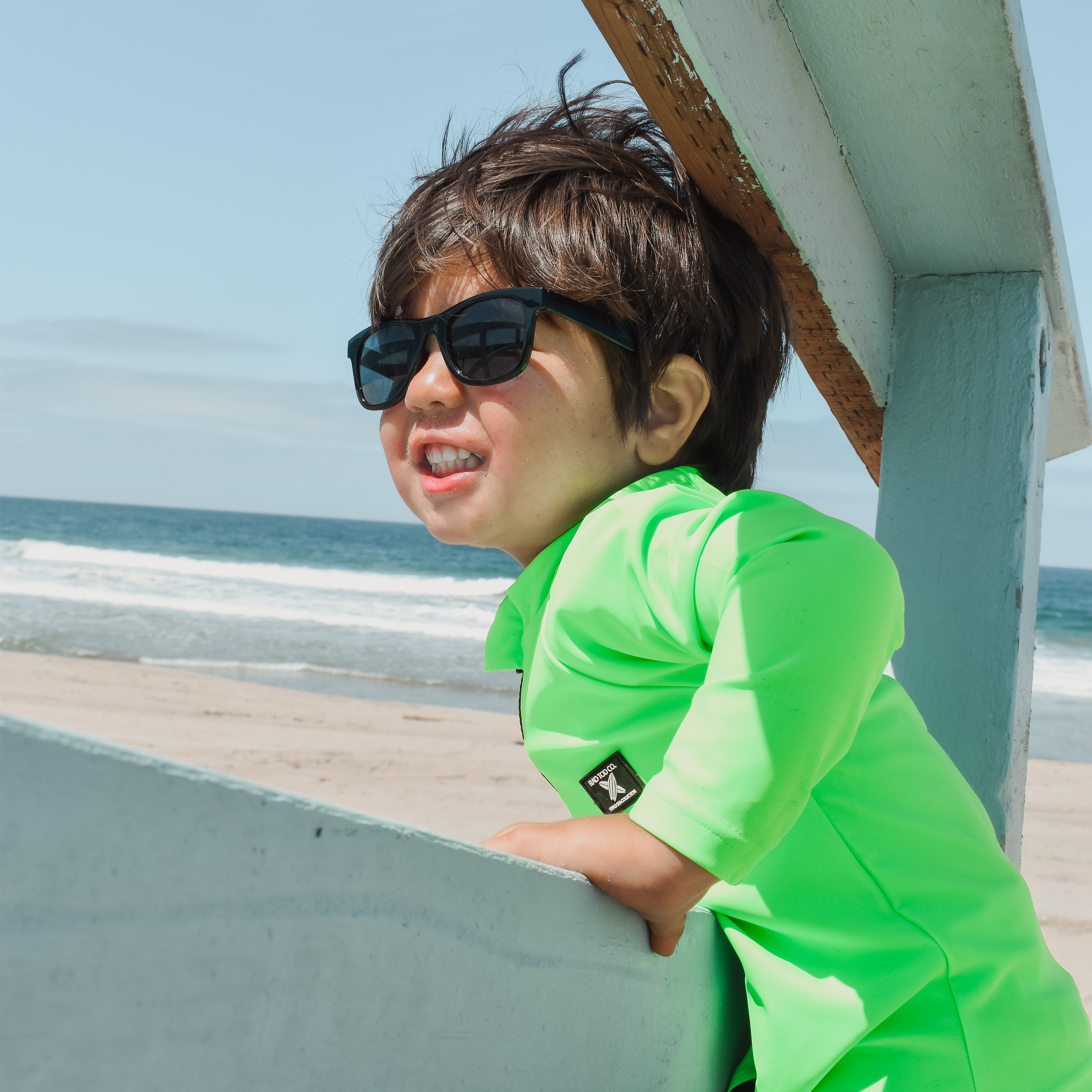 Boy smiling while wearing bright neon zip-front rashguard, protected from sun while easily visible against sandy beach background.