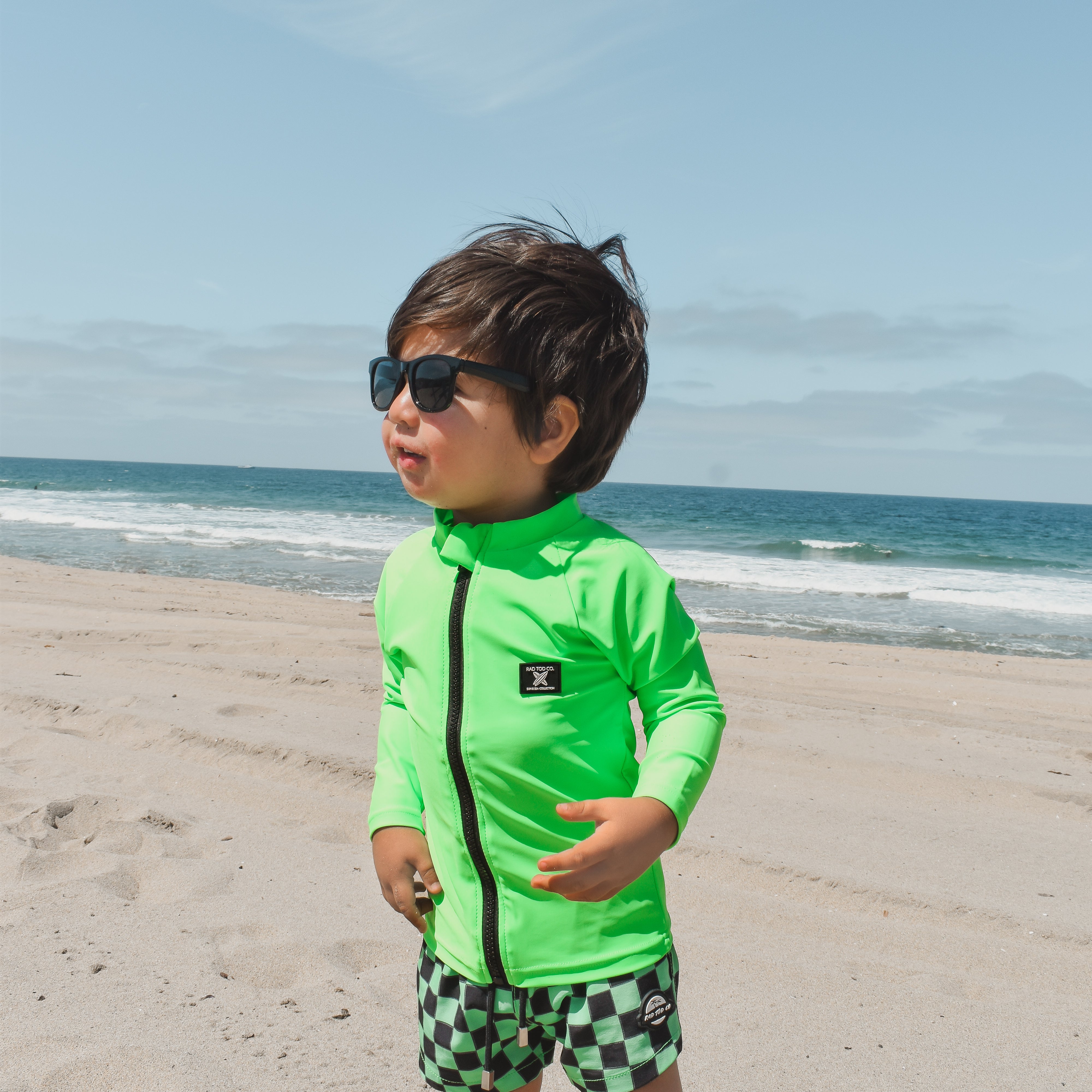 Boy hanging out in the sand wearing bright neon zip-front rashguard, protected from sun while easily visible against sandy beach background.