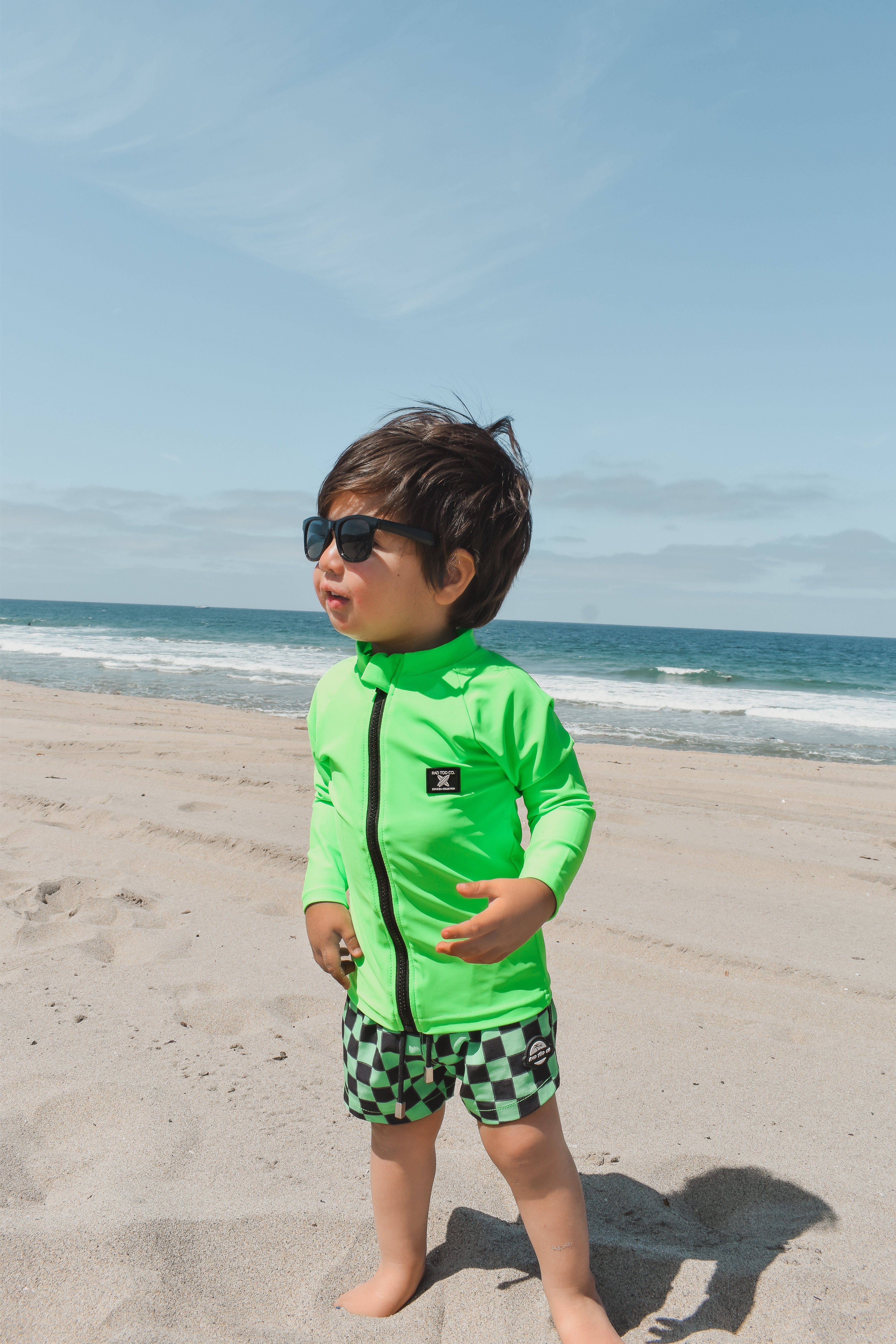 Boy hanging out in the sand wearing bright neon zip-front rashguard, protected from sun while easily visible against sandy beach background.