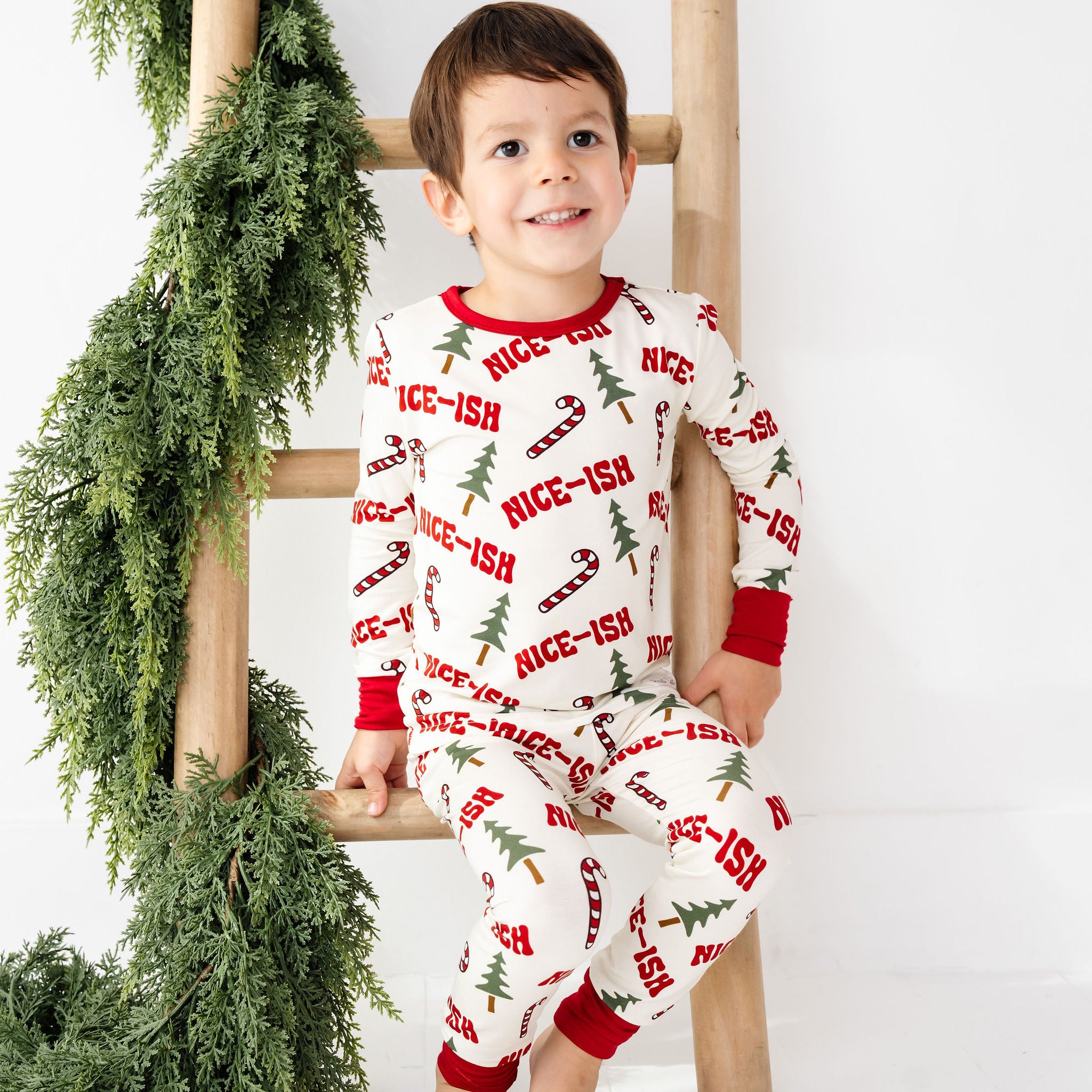 Child wearing a Christmas-themed onesie sitting on a wooden ladder with greenery.