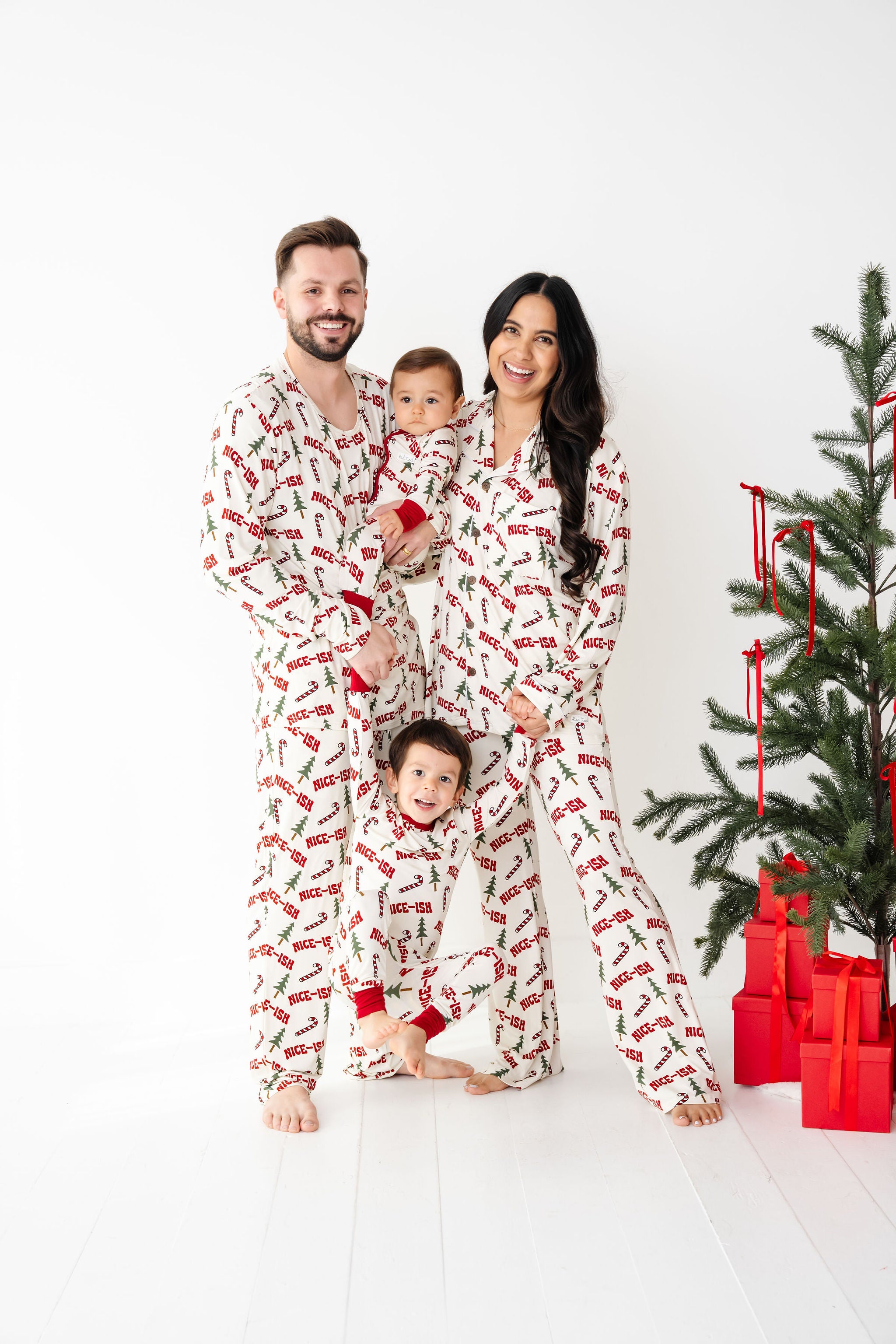 Family of four wearing matching pajamas in front of a Christmas tree with presents. They are wearing Nice-ish bamboo which is white and the writing is in RED