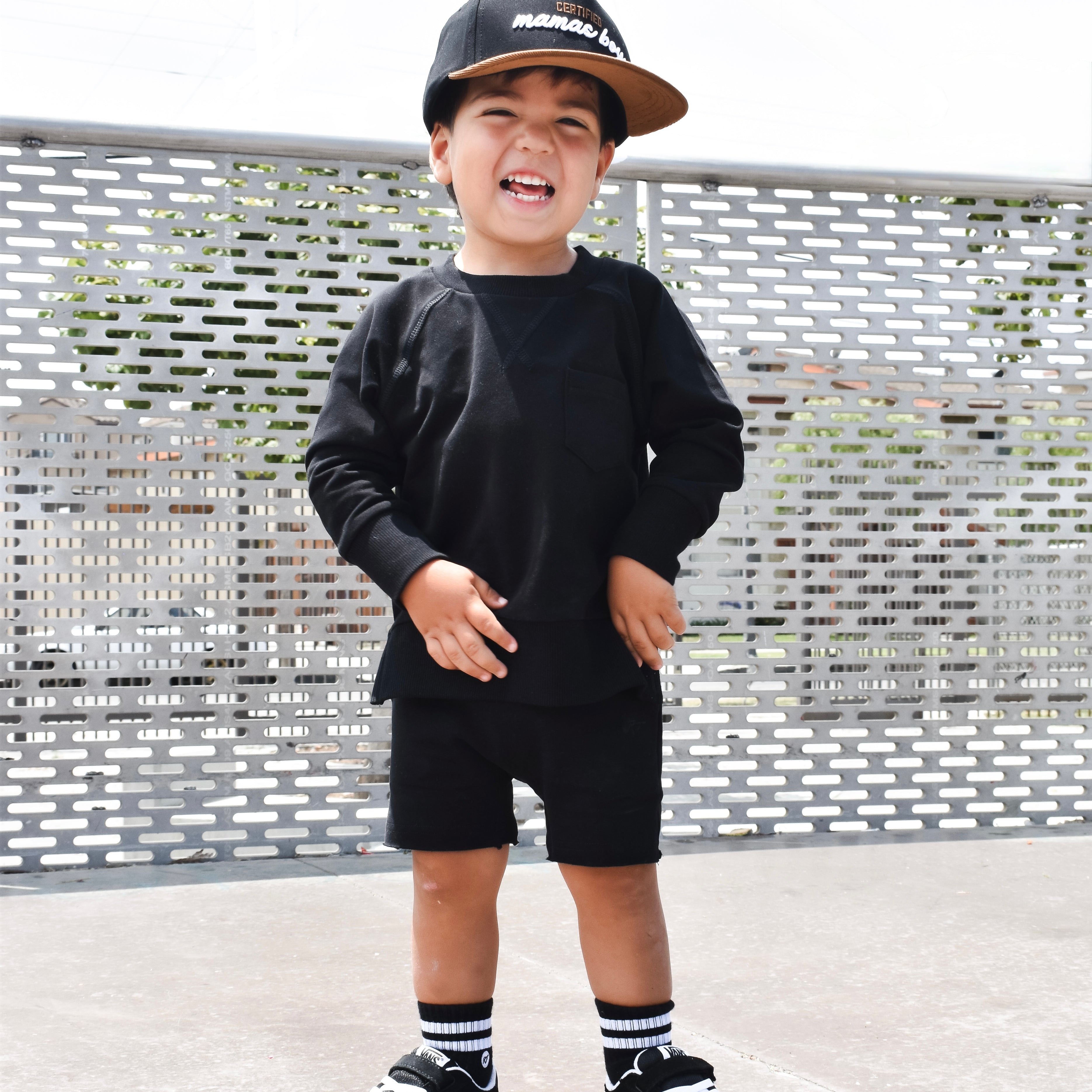 Child wearing a black outfit with a cap, standing on a concrete surface with a metal fence background.