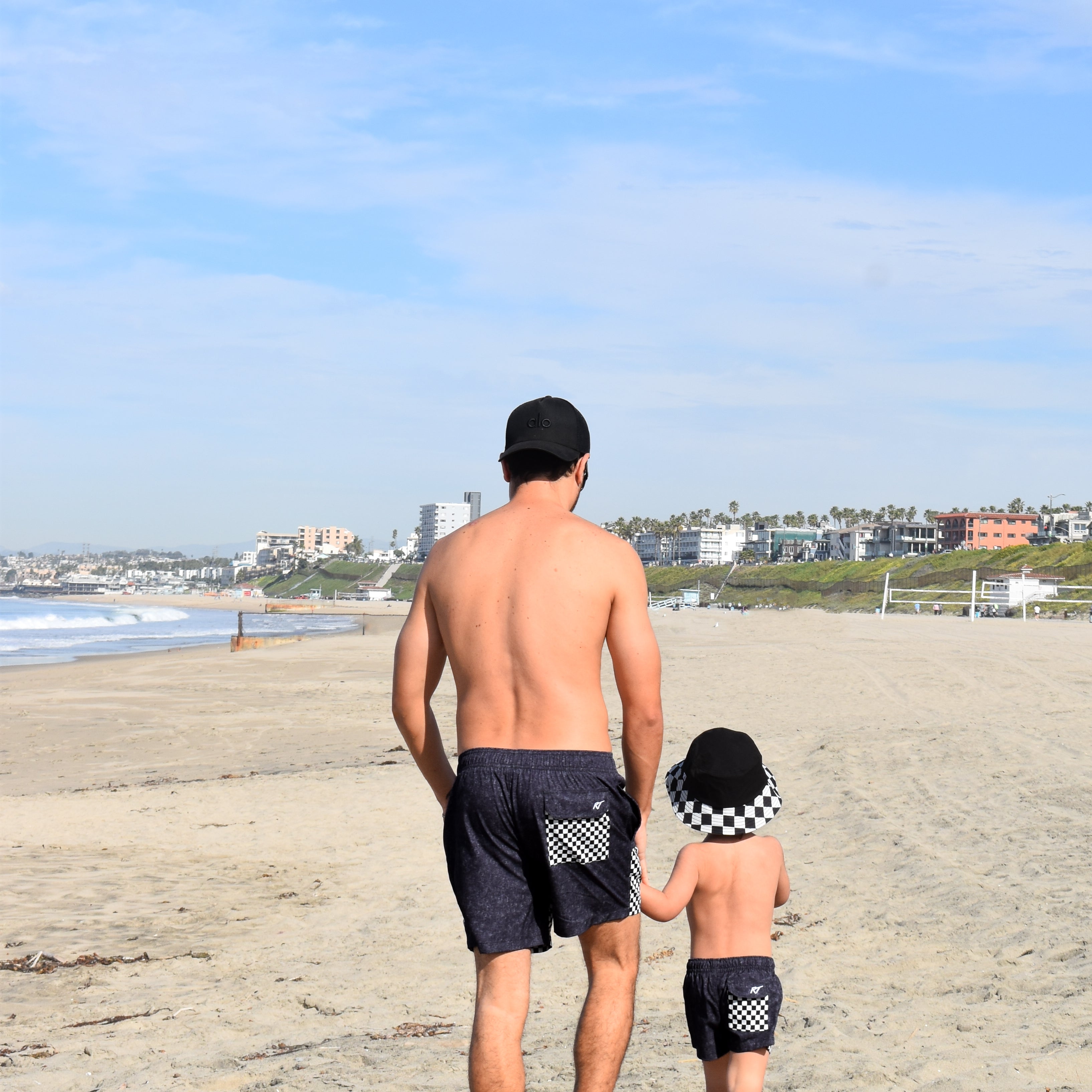 Man and child walking on a beach with a clear blue sky wearing matching board shorts