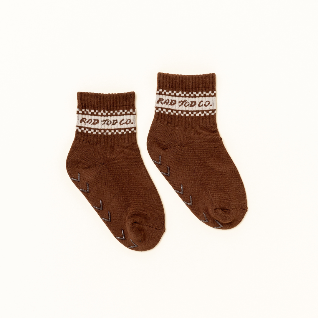 A pair of brown crew socks with a cream and brown patterned cuff featuring the text 'ROD TOD CO.' and small grip dots on the sole, shown on a plain off-white background.