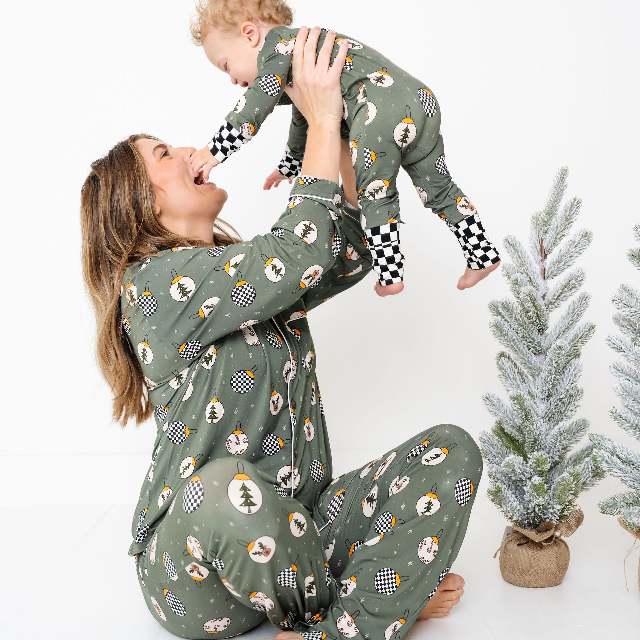 Woman and child wearing matching green pajamas with a pattern by RAD TODDLER, sitting on a white floor with small Christmas trees.