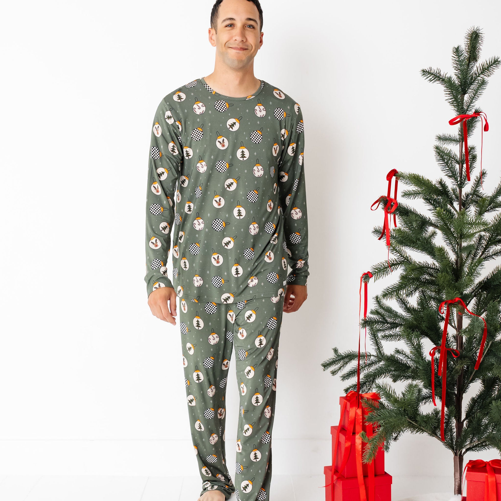 Man wearing a green patterned onesie standing next to a decorated Christmas tree with red ribbons and presents on a white background.