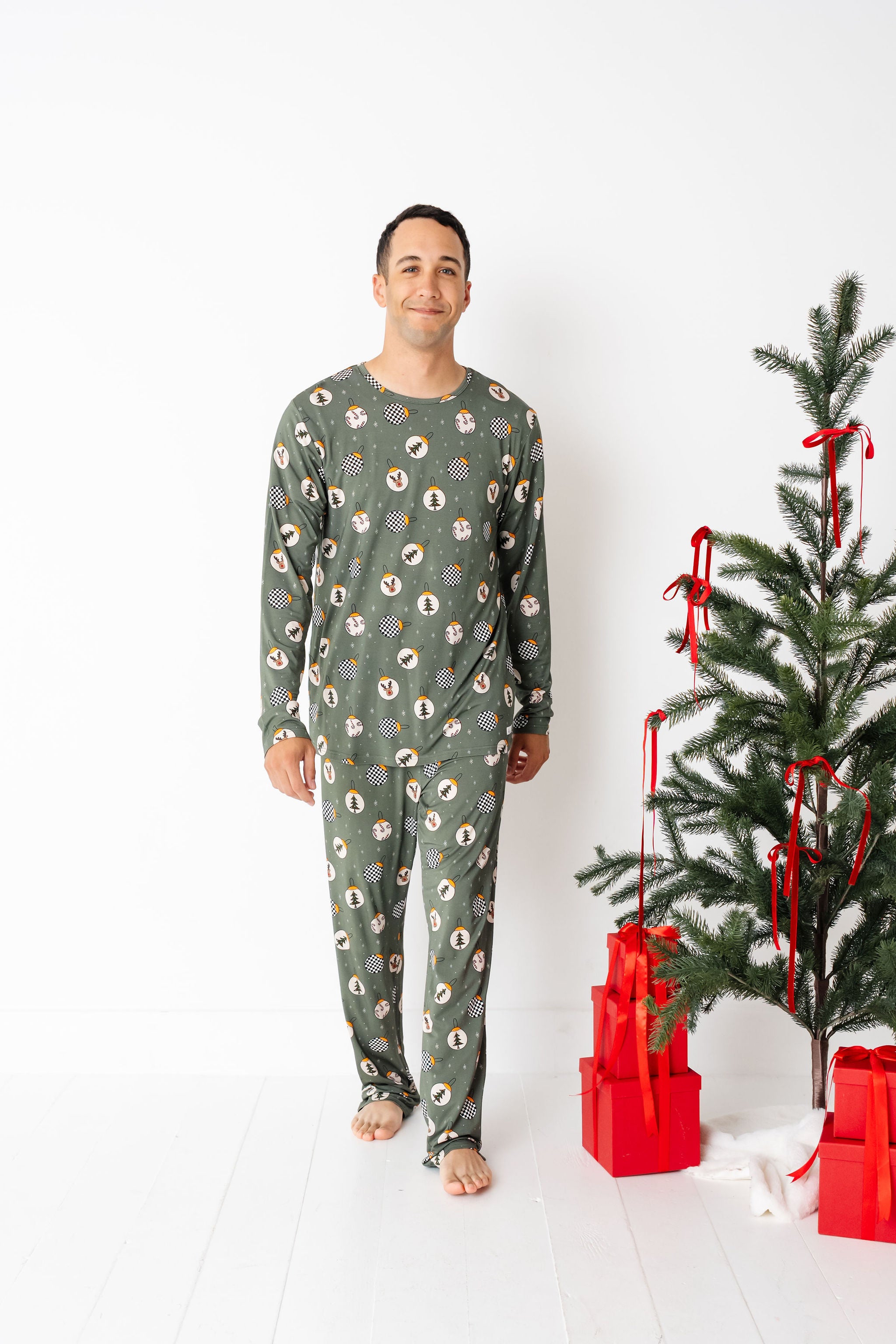 Man wearing a green patterned onesie standing next to a decorated Christmas tree with red ribbons and presents on a white background.