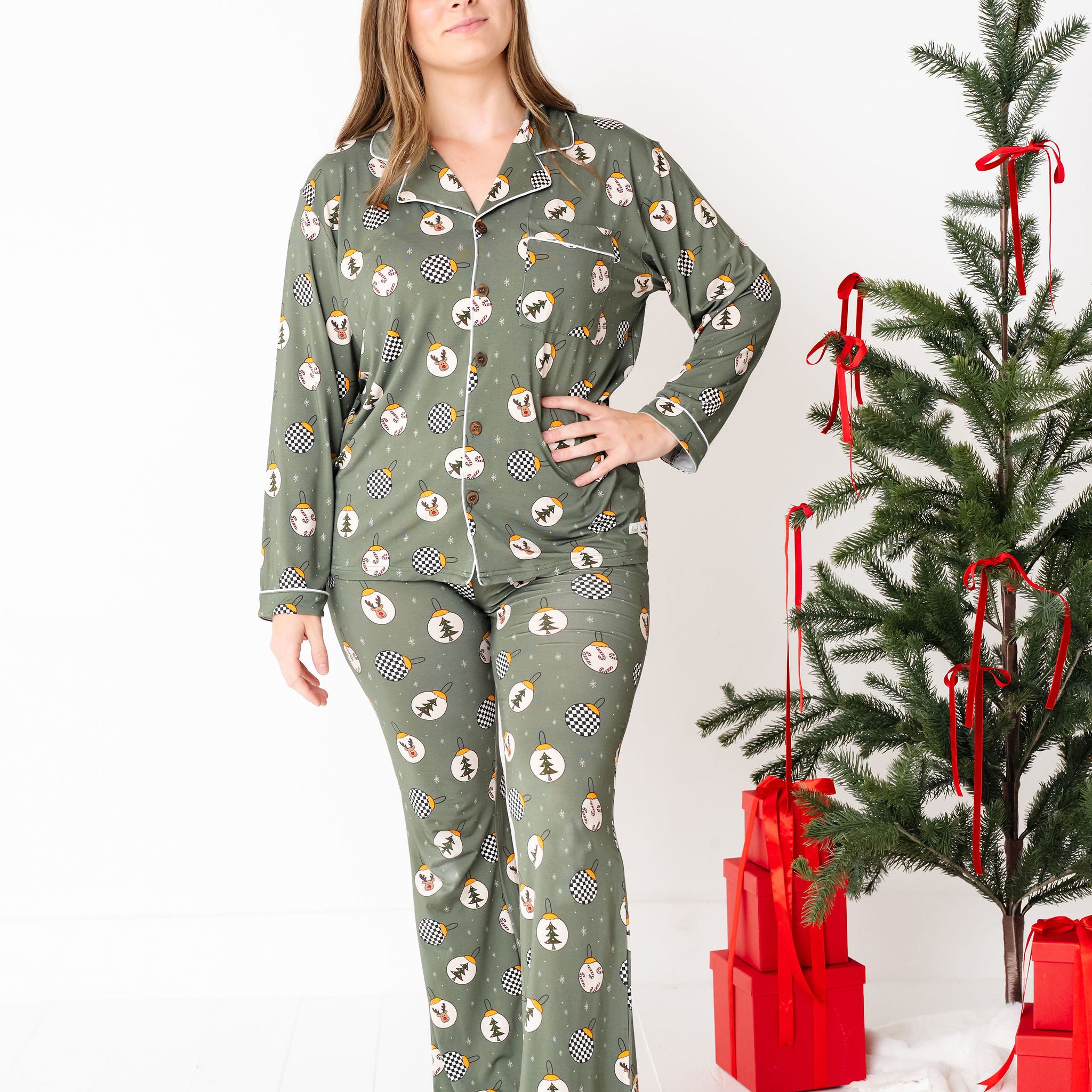 Woman wearing green patterned pajamas standing next to a decorated Christmas tree with red bows and presents.