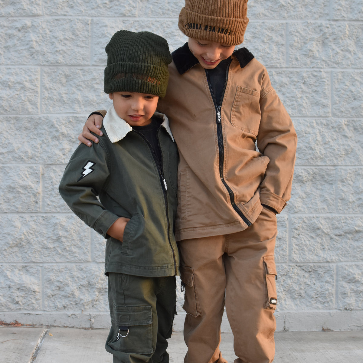 Two children standing outdoors against a light gray brick wall, wearing matching streetwear outfits. The child on the left wears an olive green jacket and pants with a dark green beanie, and the child on the right wears a tan/brown jacket and matching pants with a brown beanie.