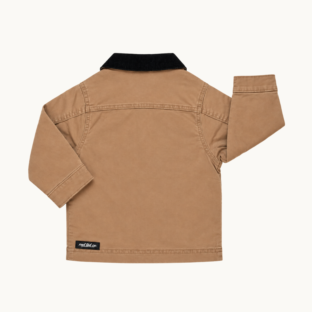 A tan long-sleeve utility jacket shown from the back, featuring a black corduroy collar and a small black brand patch near the bottom left hem, set against a plain off-white background.