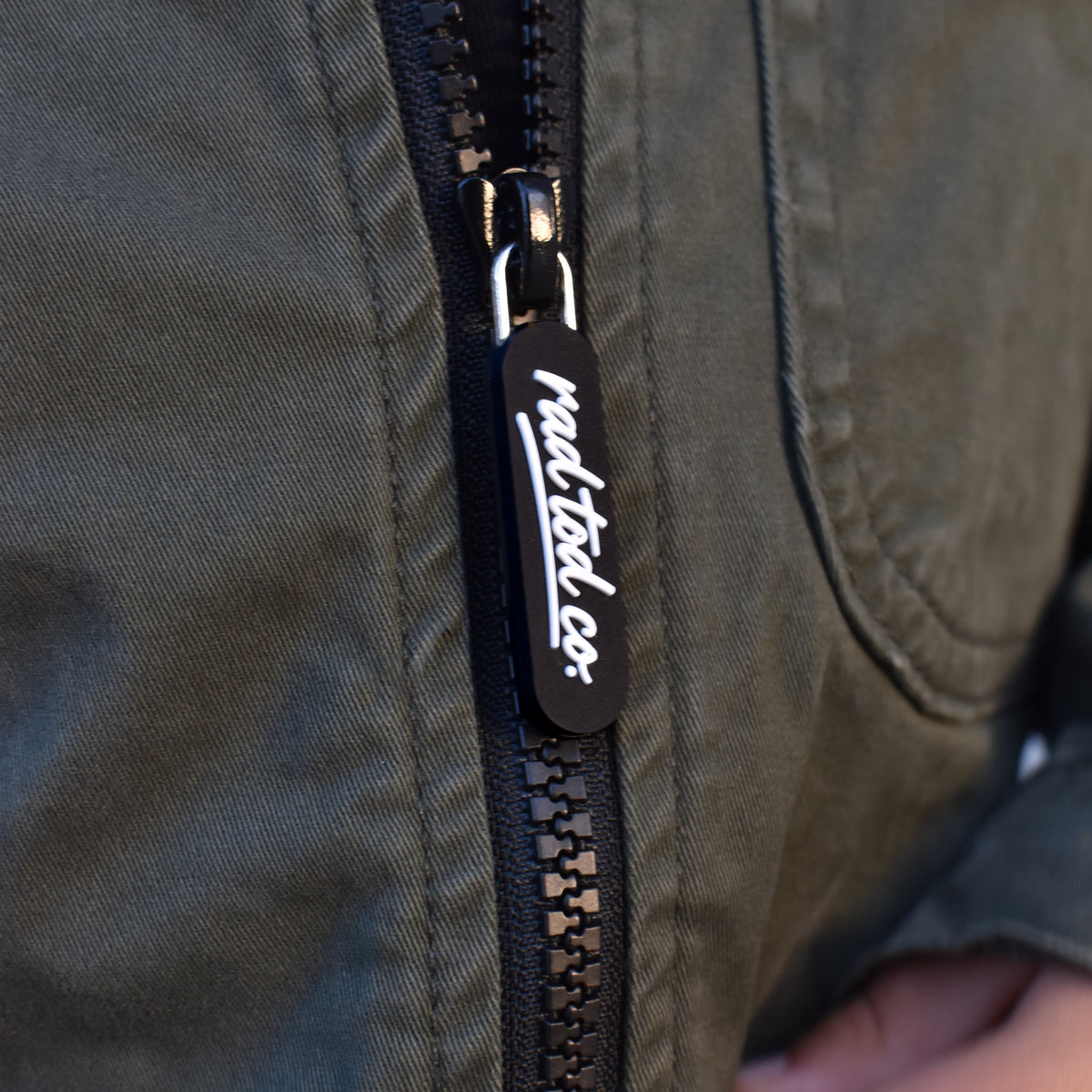 Close-up of a black zipper on an olive green jacket, featuring a black pull tab with white script text reading 'RAD TOD CO.' and a hand holding the zipper open.