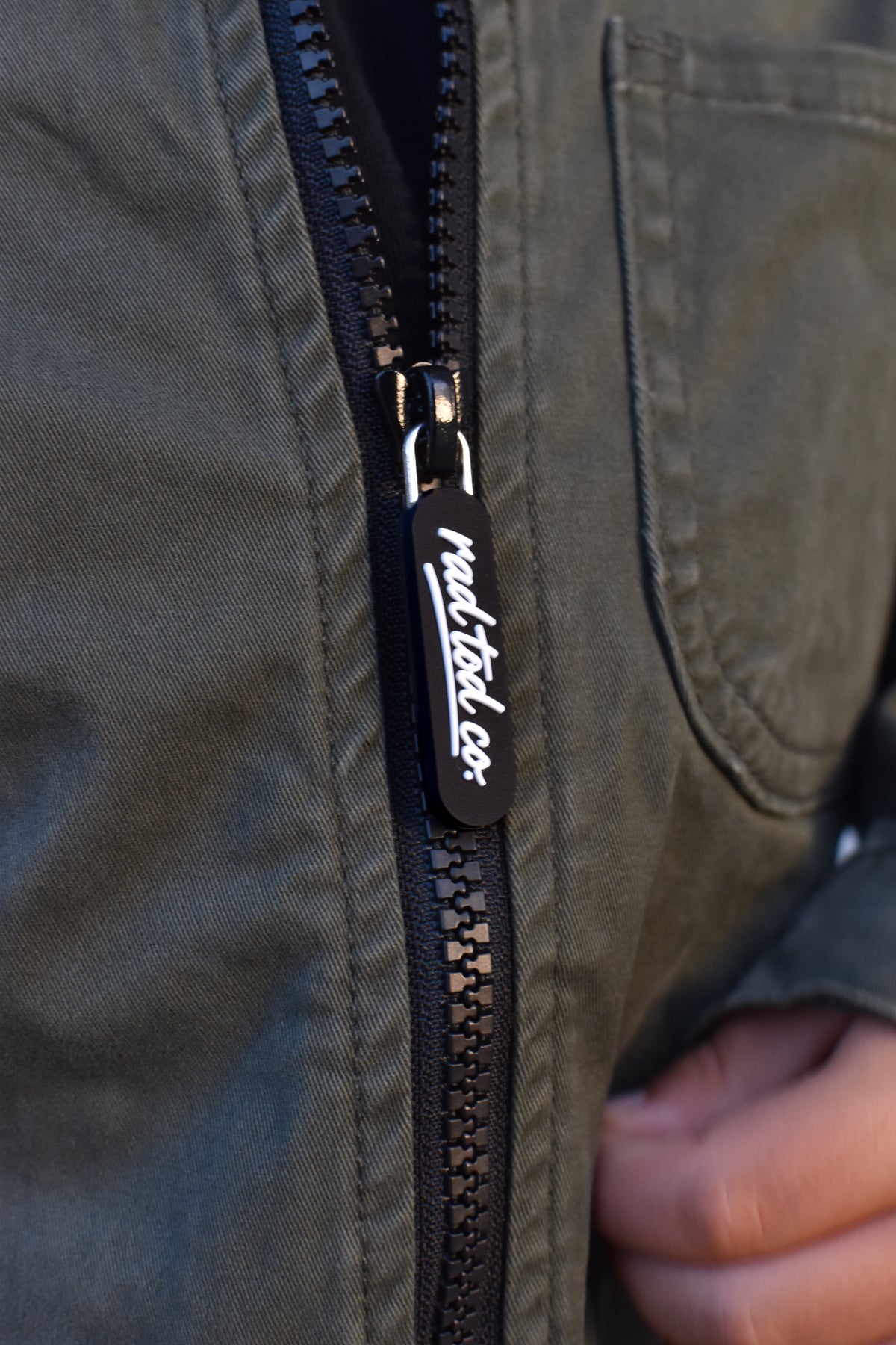 Close-up of a black zipper on an olive green jacket, featuring a black pull tab with white script text reading 'RAD TOD CO.' and a hand holding the zipper open.