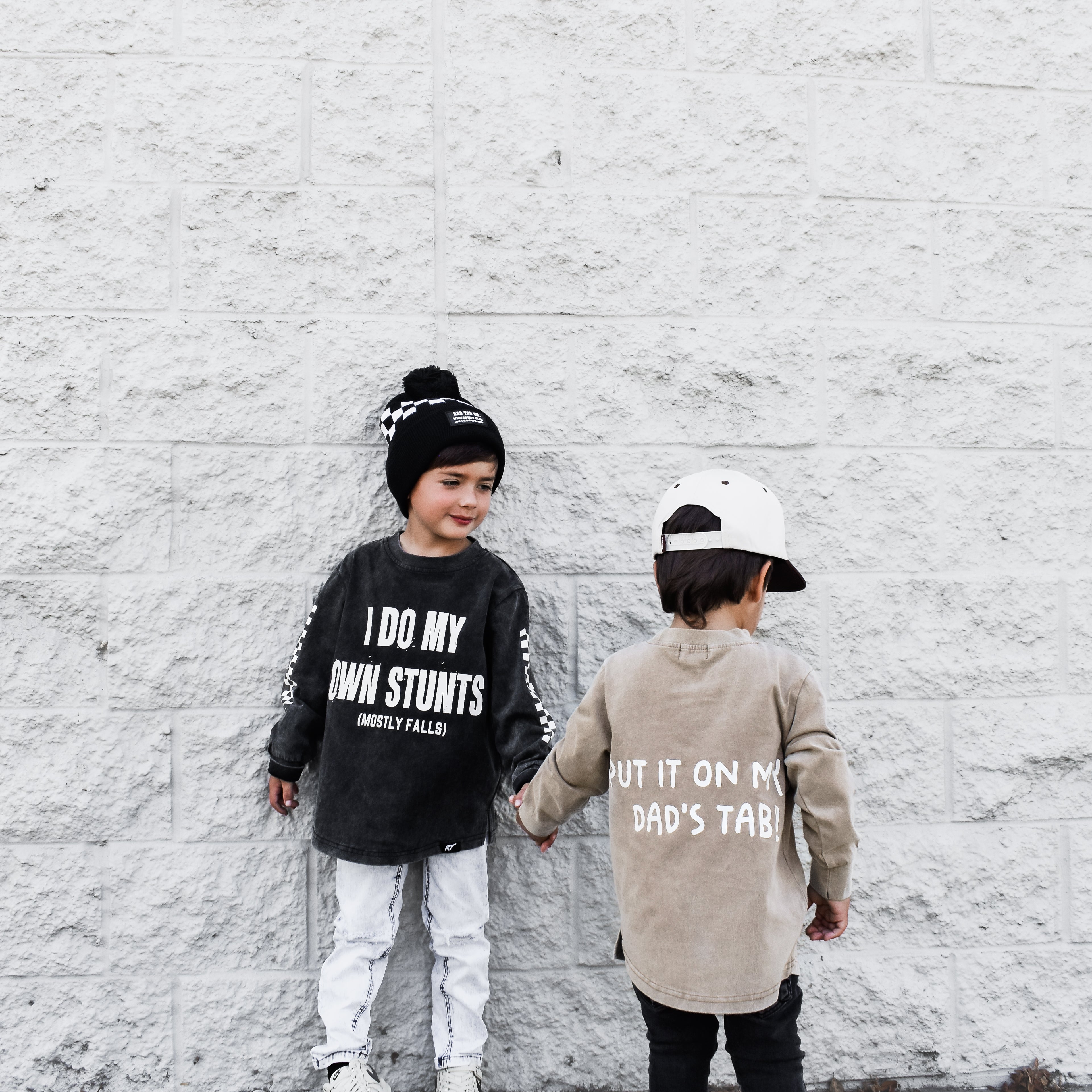 Two children standing against a white brick wall, wearing Rad Toddler t-shirts with text.