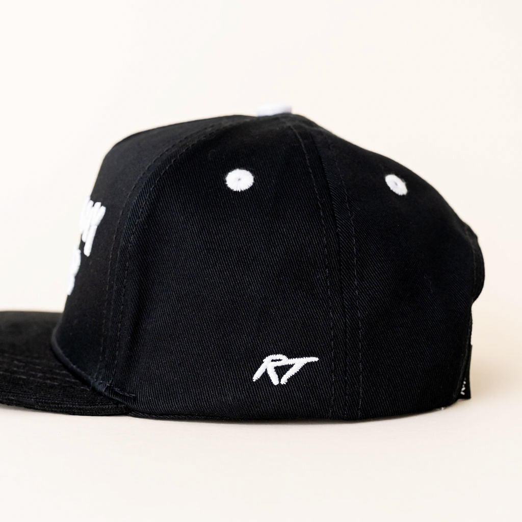 A black snapback cap shown from a side angle, featuring a white embroidered stylized logo on the front side panel and a matching white logo on the brim, set against a plain off-white background.