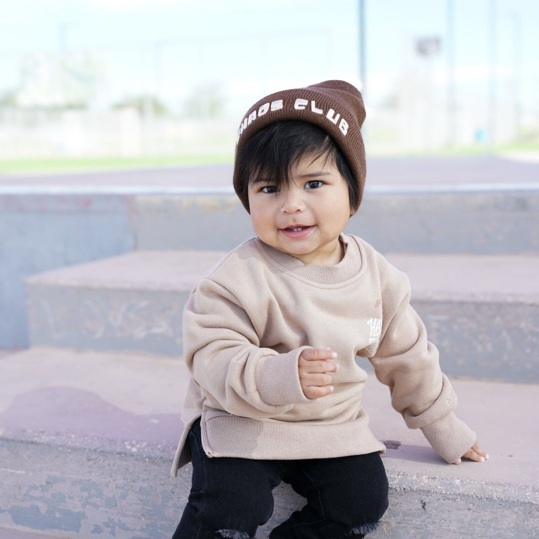 Child wearing a beige sweatshirt, black pants, and a brown beanie sitting on steps.
