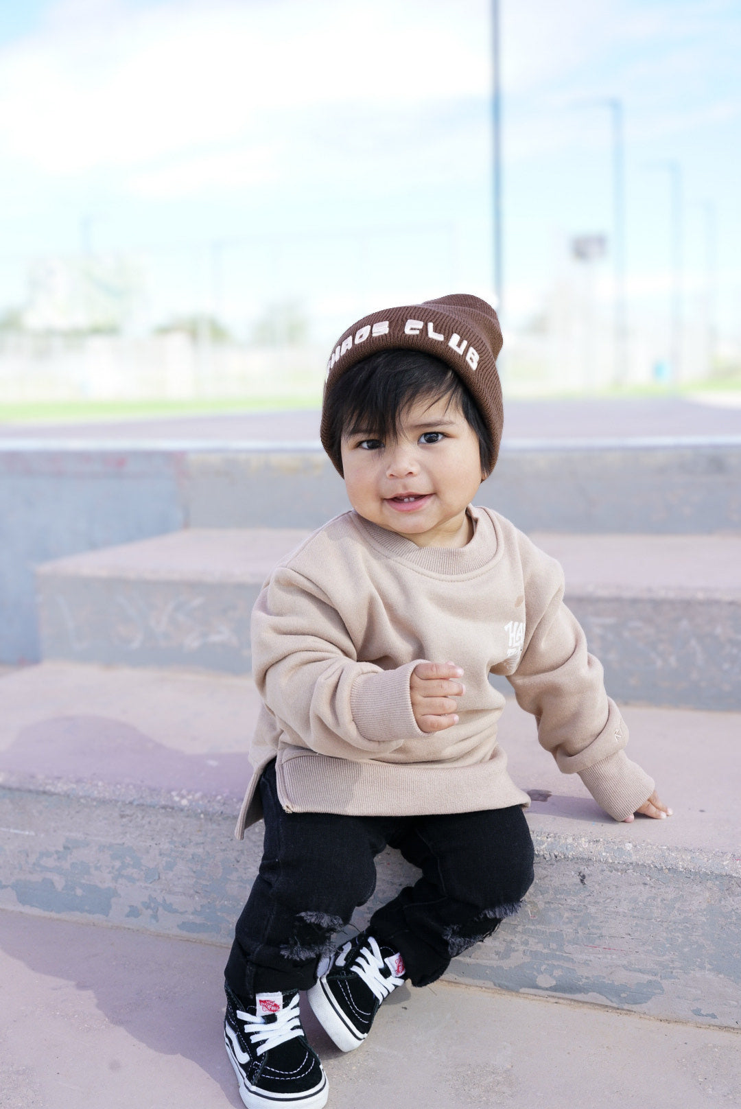 Child wearing a beige sweatshirt, black pants, and a brown beanie sitting on steps.