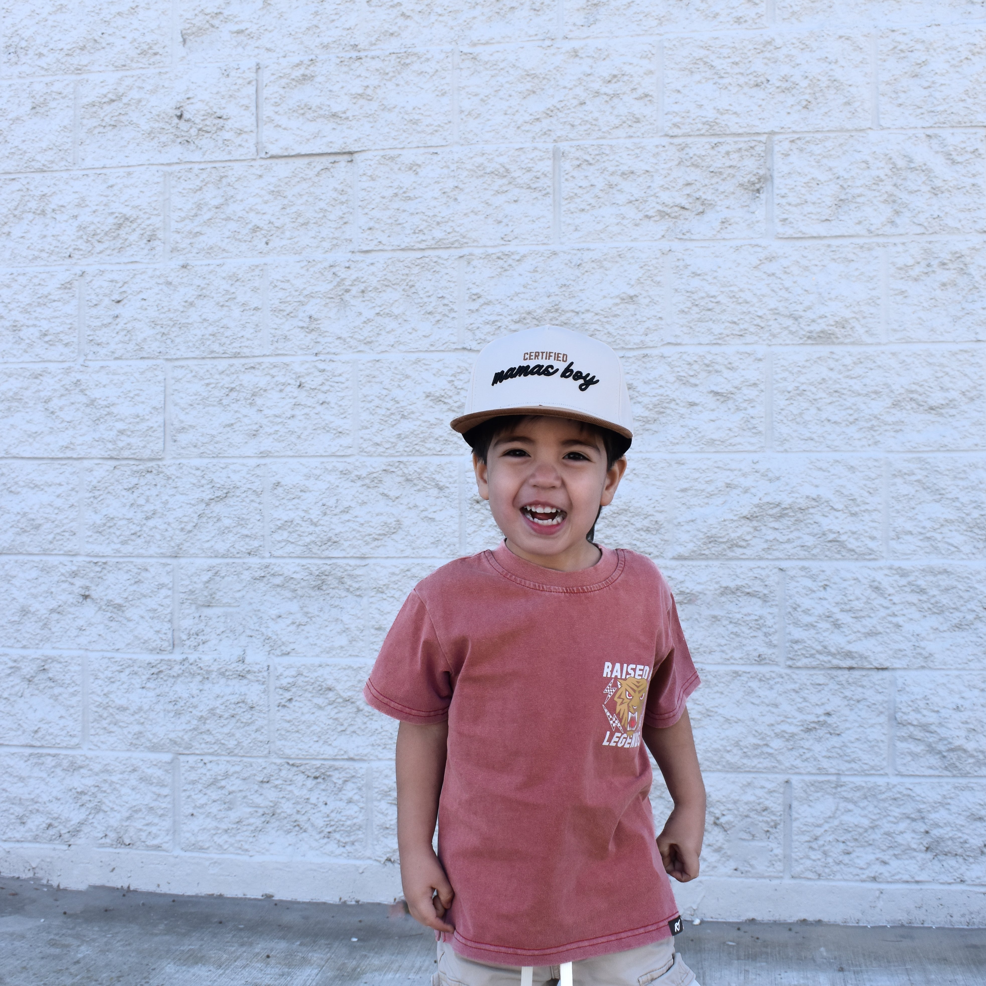 Child wearing a red shirt and beige cap standing against a white brick wall.
