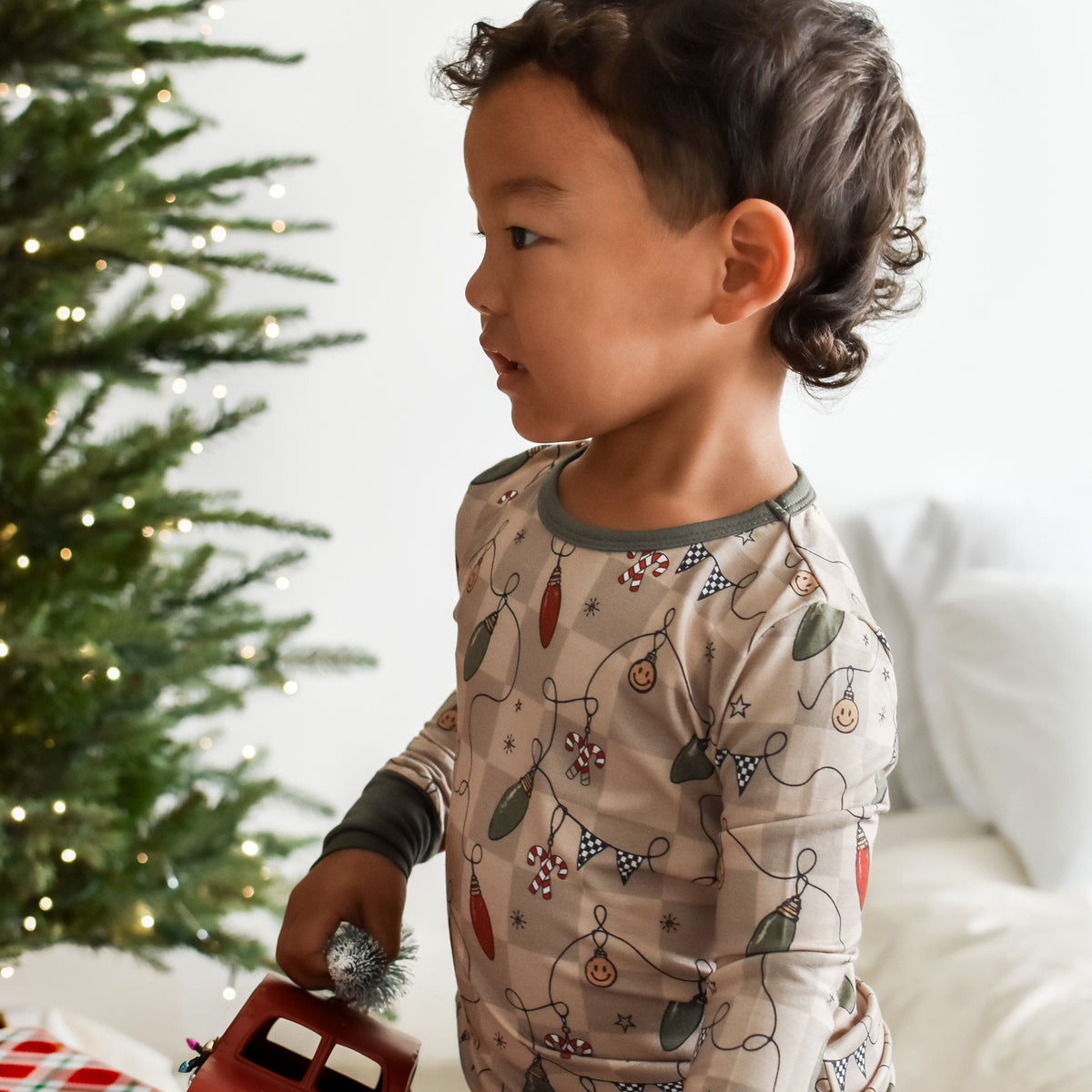 A toddler wearing a holiday-themed long-sleeve pajama set with a Christmas tree and ornament print, standing next to a decorated Christmas tree and holding a small red toy truck.