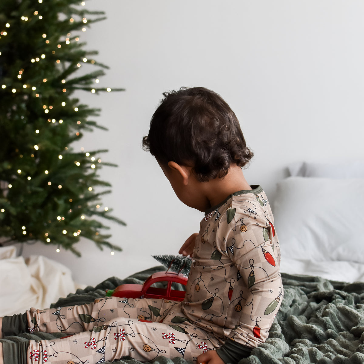 A toddler wearing a beige Christmas-themed long-sleeve pajama set with a holiday lights print, sitting on a dark green blanket next to a decorated Christmas tree.
