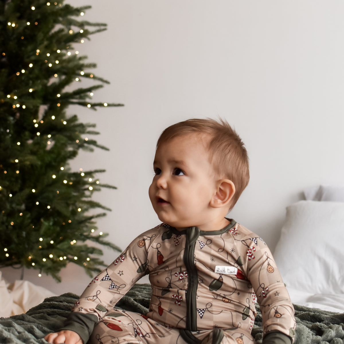 A baby sitting on a textured green blanket next to a lit Christmas tree, wearing a beige holiday-themed long-sleeve sleeper with a dark zipper.