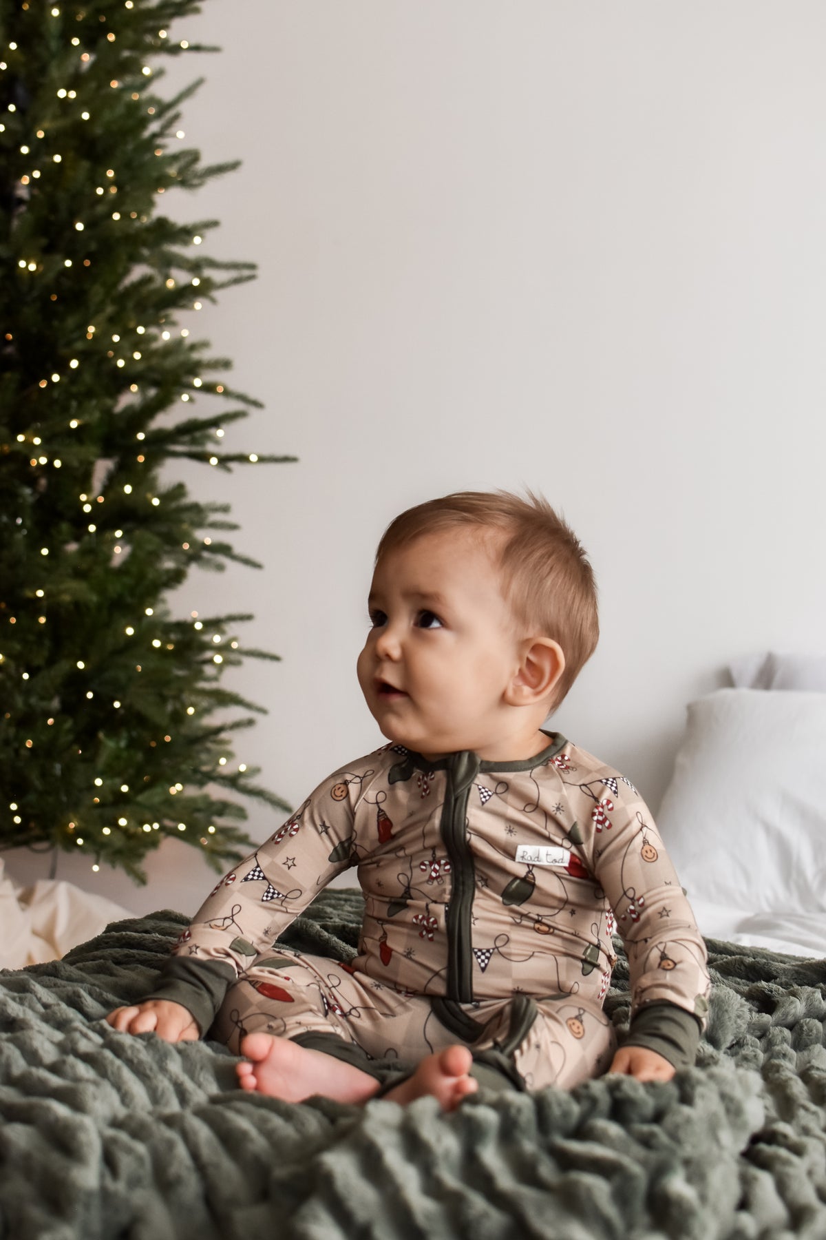 A baby sitting on a textured green blanket next to a lit Christmas tree, wearing a beige holiday-themed long-sleeve sleeper with a dark zipper.