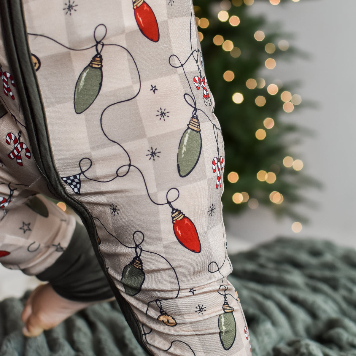 A close-up of a person wearing holiday-themed pajama pants with a Christmas light pattern, standing on a textured gray blanket with a blurred Christmas tree in the background.
