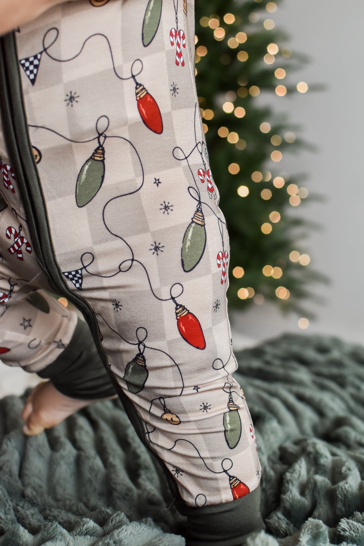 A close-up of a person wearing holiday-themed pajama pants with a Christmas light pattern, standing on a textured gray blanket with a blurred Christmas tree in the background.