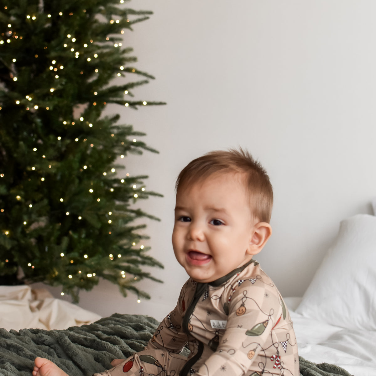 A baby sitting on a green blanket in front of a Christmas tree, wearing a beige long-sleeve pajama with a winter botanical pattern.