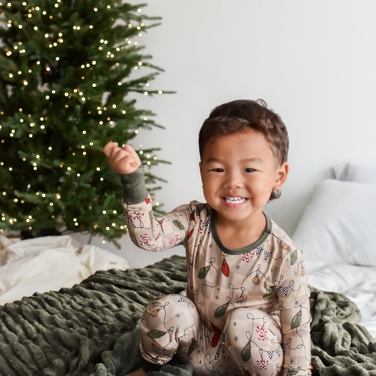 A smiling young child wearing a festive holiday pajama set sitting on a ribbed dark green blanket next to a decorated Christmas tree.
