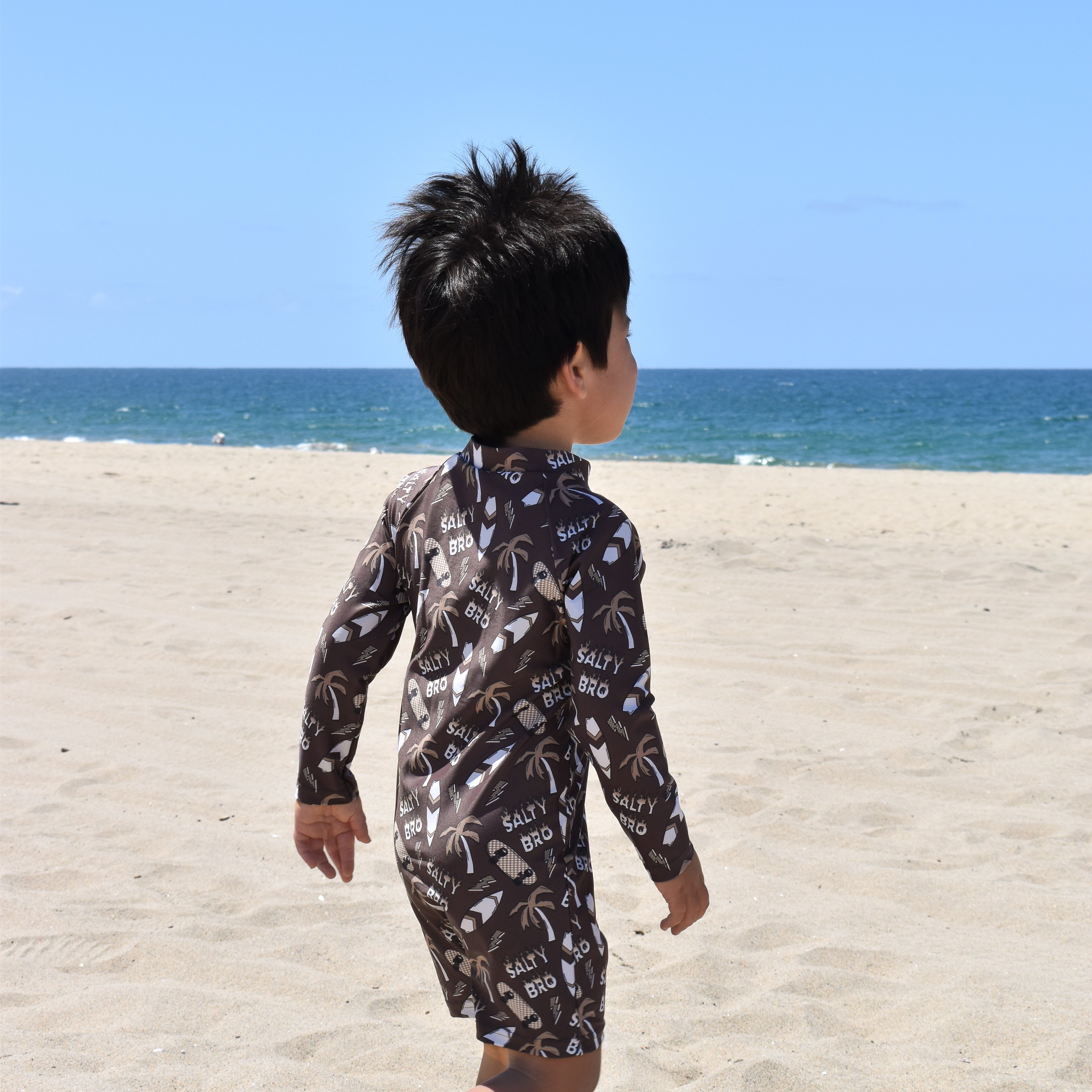Child in a patterned swimsuit walking on a sandy beach with clear blue sky and ocean.
