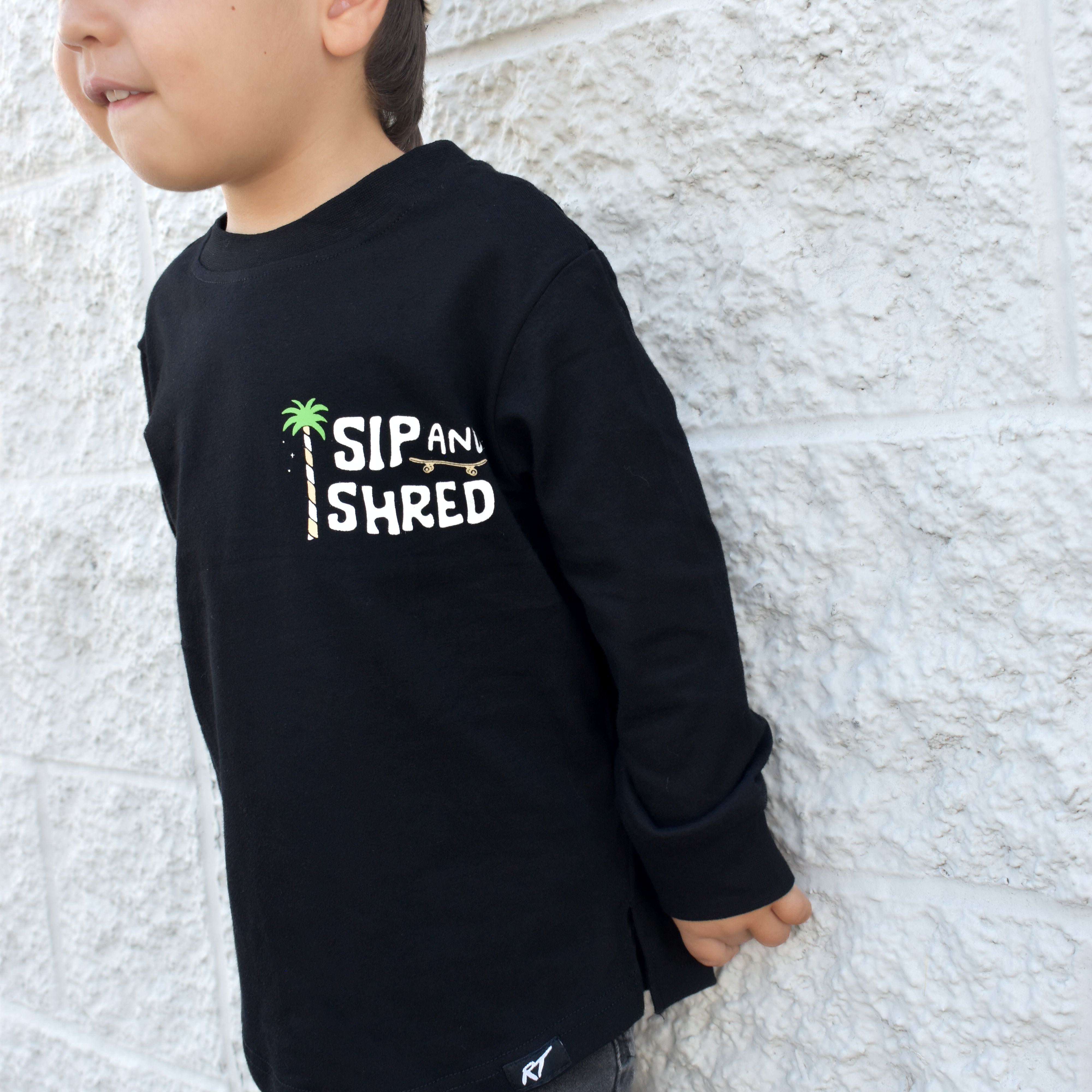 Child wearing a black sweatshirt with 'Sip and Shred' text against a light stone wall.