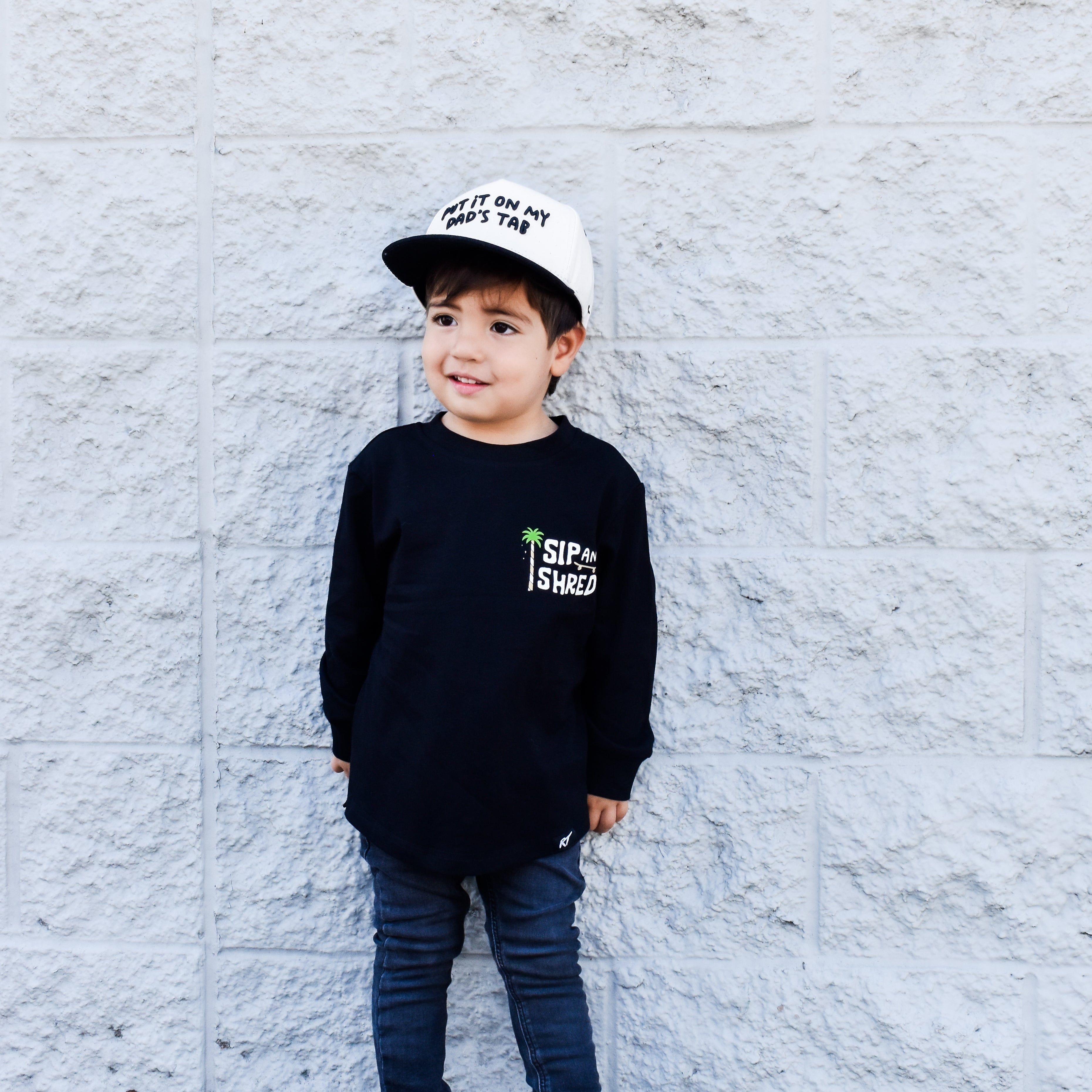 Child wearing a black sweatshirt with text, blue jeans, and a white cap against a light gray brick wall.