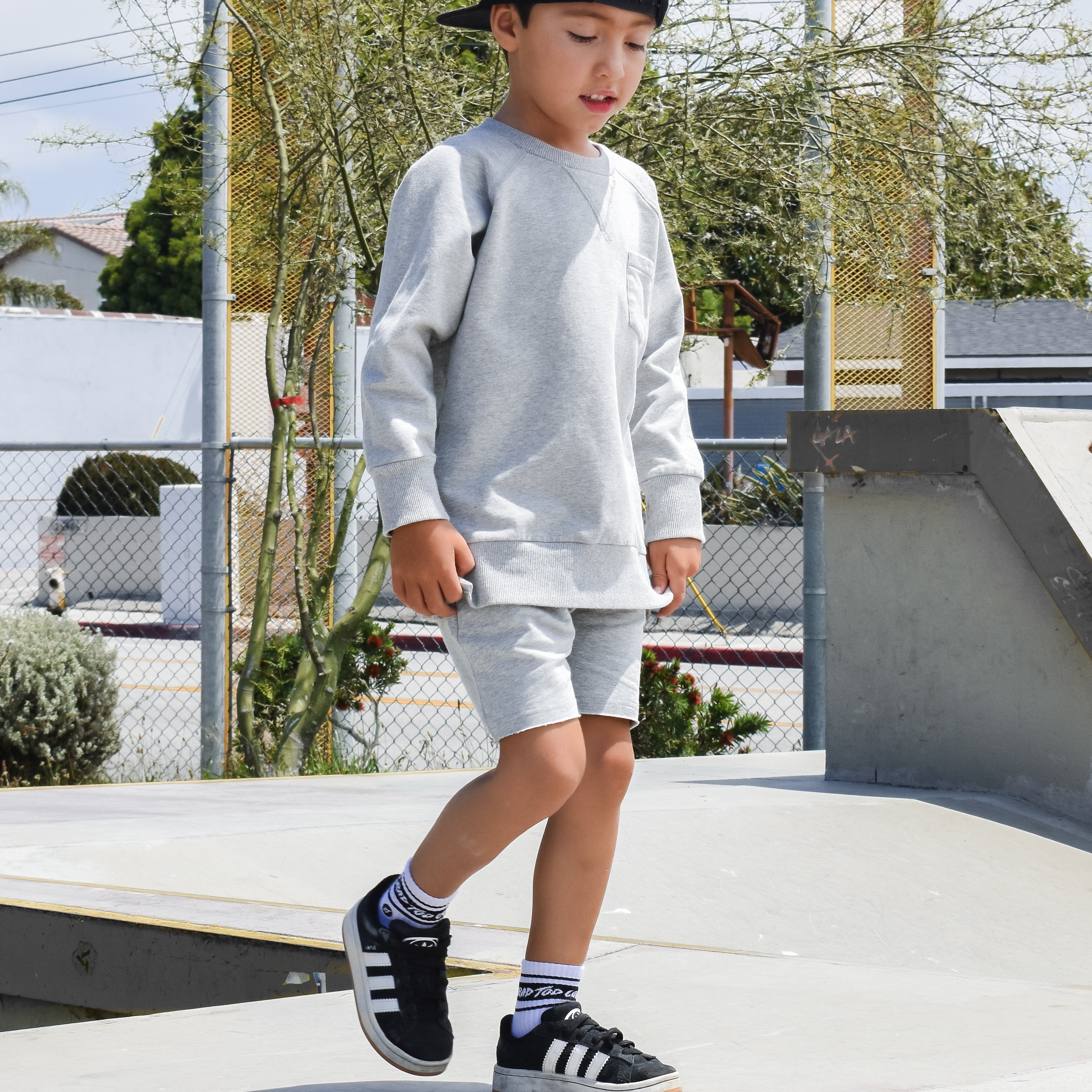 Child in gray sweatshirt and shorts walking on a concrete surface with trees and buildings in the background