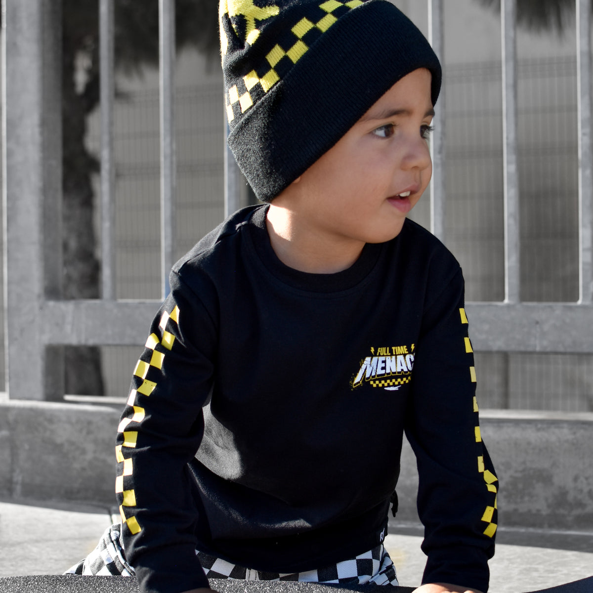 A child wearing a black and yellow checkered beanie and a black long-sleeve shirt with yellow accents sits on a skateboard in front of a gray metal fence.