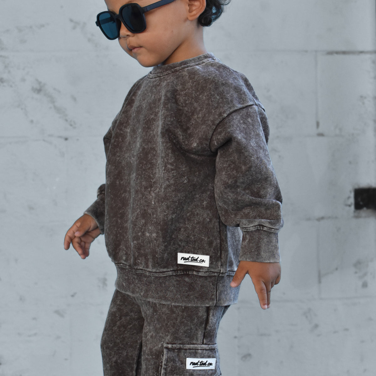 A toddler wearing a matching washed dark brown sweatshirt and cargo pants set with sunglasses, standing against a light gray concrete wall.